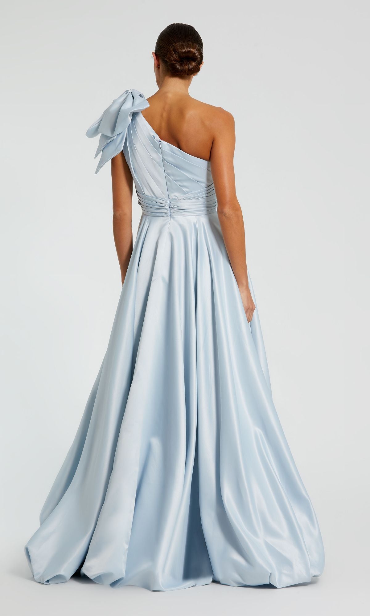 Woman wearing the Mac Duggal 11800 Bow Long Bubble Prom Dress, a light blue one-shoulder A-line evening gown with a large bow on the shoulder, stands with her back to the camera.