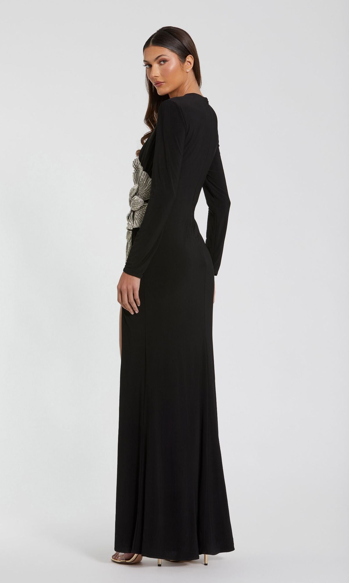 A woman stands in a three-quarter pose against a plain background, modeling the Mac Duggal 11809 long sleeve black formal dress with silver side detailing and a high slit.