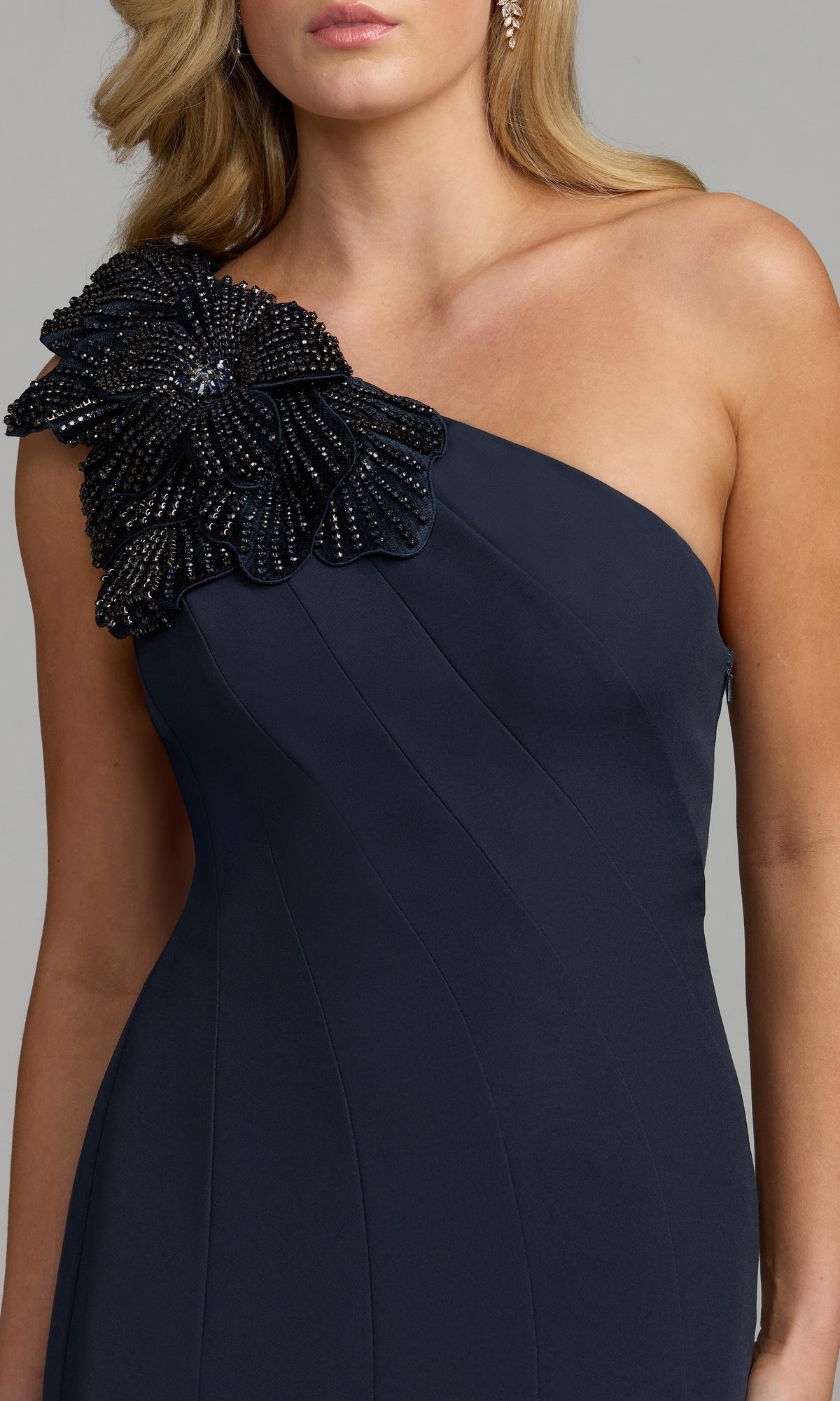 A woman wearing the Mac Duggal 11810 Beaded-One-Shoulder Prom Gown in navy, which features a striking one-shoulder design adorned with a large, beaded floral embellishment on the shoulder.