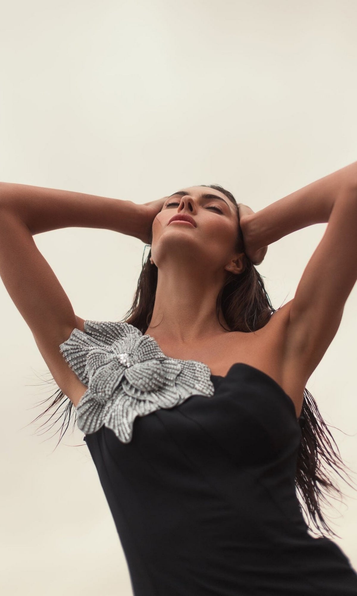 A woman poses outdoors in the Mac Duggal 11810 Beaded-One-Shoulder Prom Gown, a black formal dress featuring a large silver flower detail, with her hands behind her head and gazing upward against a neutral sky.