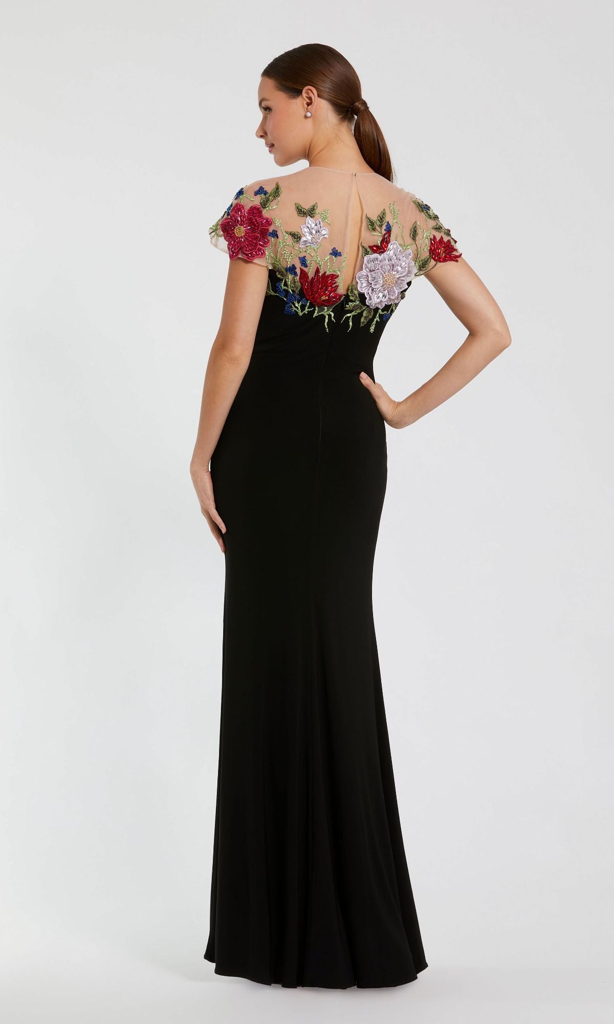 A woman stands with her back to the camera, one hand on her hip, wearing the Mac Duggal 11817 Floral Long Black Formal Dress featuring a sheer, floral-embroidered top.
