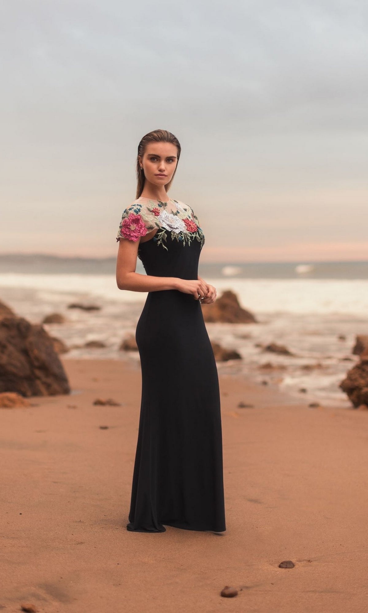 Wearing the Mac Duggal 11817 Floral Long Black Formal Dress, a woman stands on a sandy beach as her embroidered gown flows in the sunset by the ocean and rocky shore.