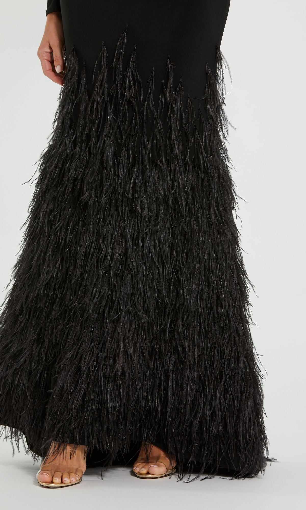 A person in a Mac Duggal 11839 Feather Long Black Formal Dress with a feathered skirt and open-toe nude heels stands on a light-colored floor.