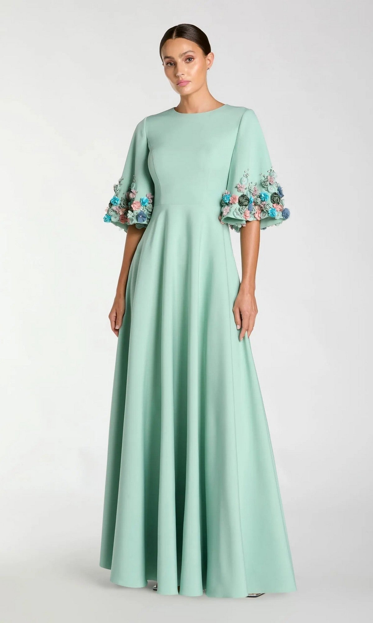 A woman in the Mac Duggal 11854 Bell-Sleeve Long Formal Dress—a pastel green, floor-length evening gown with multicolored floral embellishments—stands against a plain background.