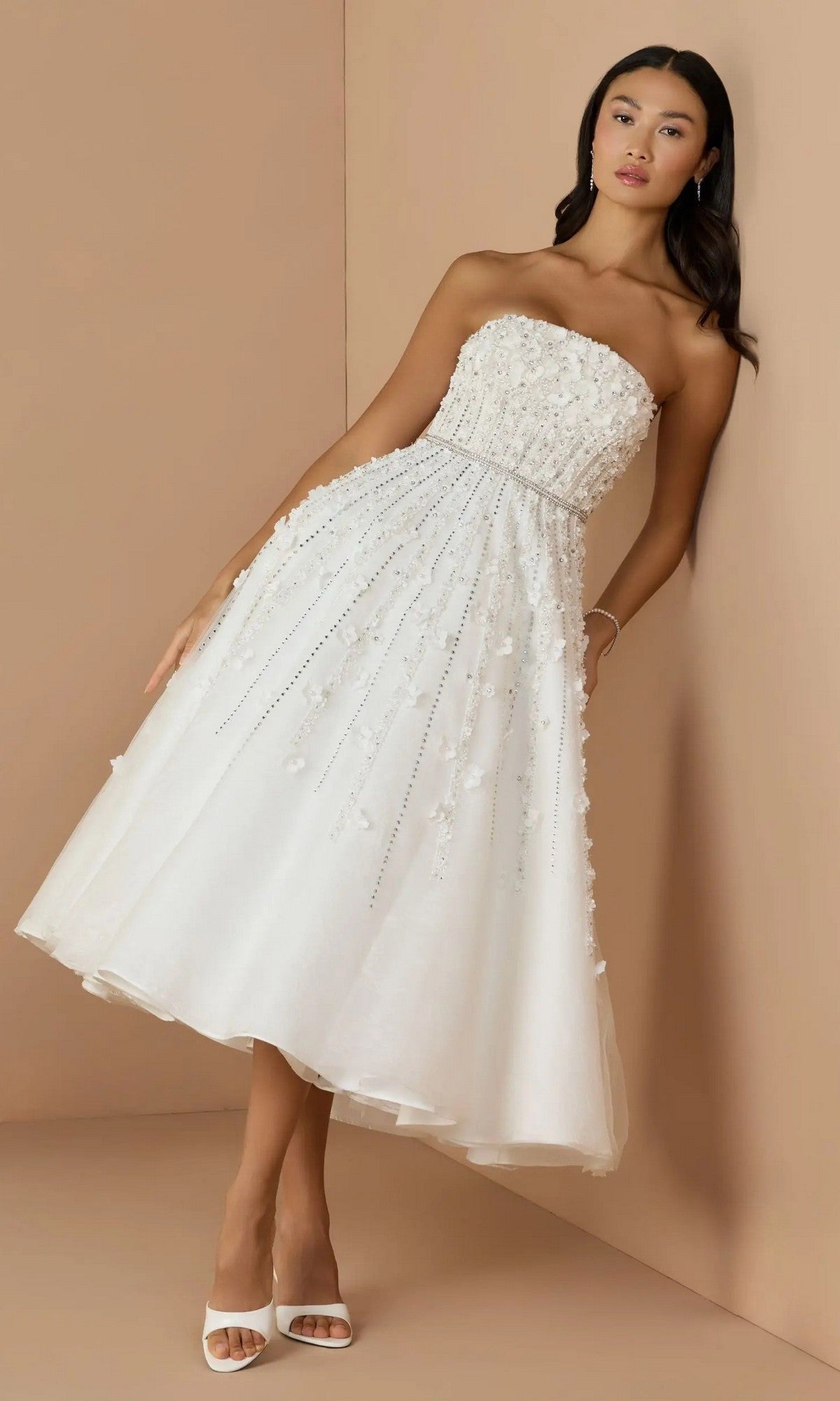 A woman in the Mac Duggal 11865 strapless champagne party dress, featuring floral embellishments and beading, stands against a beige wall wearing white heels.