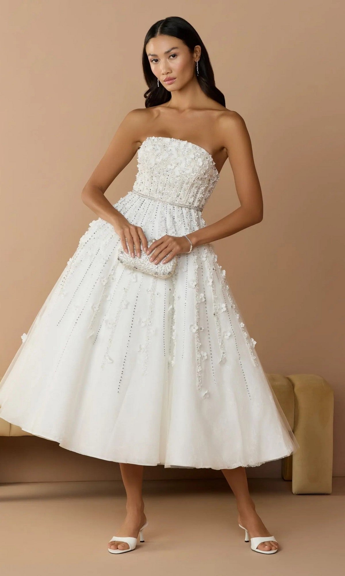 A woman wears the Mac Duggal 11865 Strapless Ivory Party Dress, featuring floral embellishments, paired with a matching clutch and white heels, as she poses against a beige background.