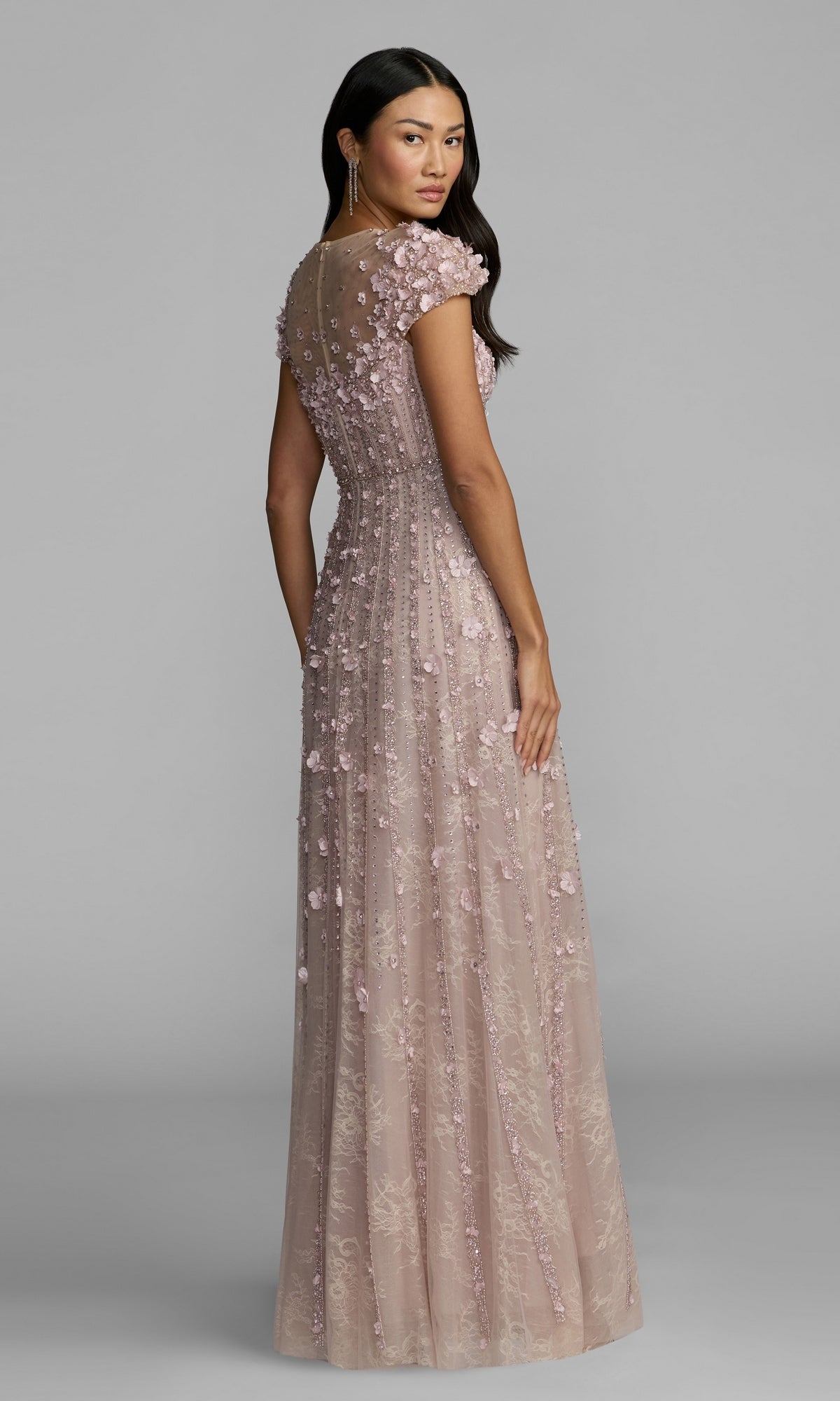 A woman wearing the Long Beaded Lilac Formal Dress—Mac Duggal 11866, featuring a floor-length skirt, short sleeves, floral appliqué, a sheer upper back, and a flowing silhouette, stands against a plain gray background.