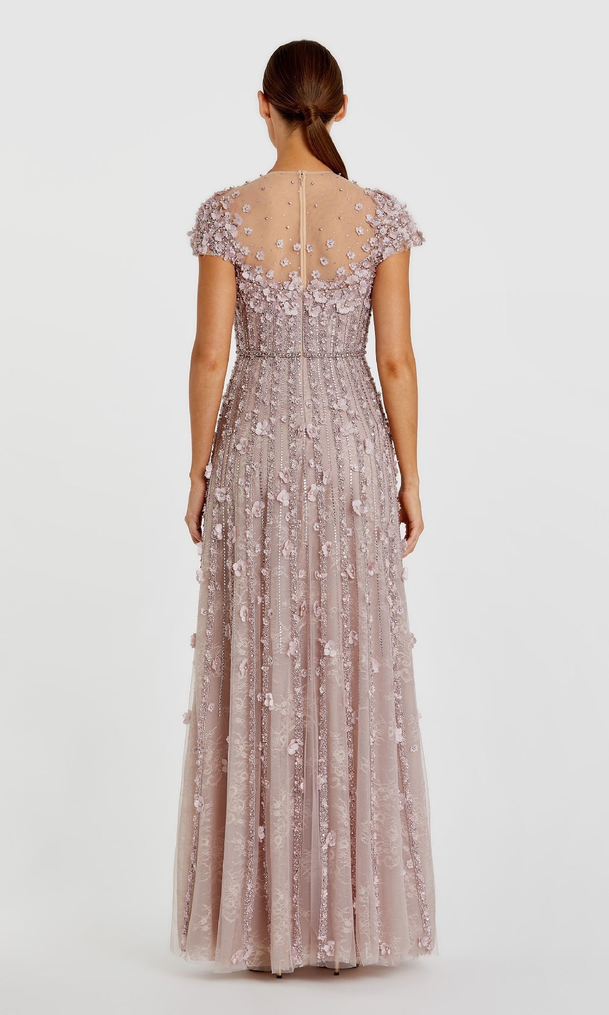 A woman with brown hair in a low ponytail models the Mac Duggal 11866 Long Beaded Lilac Formal Dress, a floor-length gown with sheer short sleeves and 3-D floral appliques, shown from the back against a plain background.