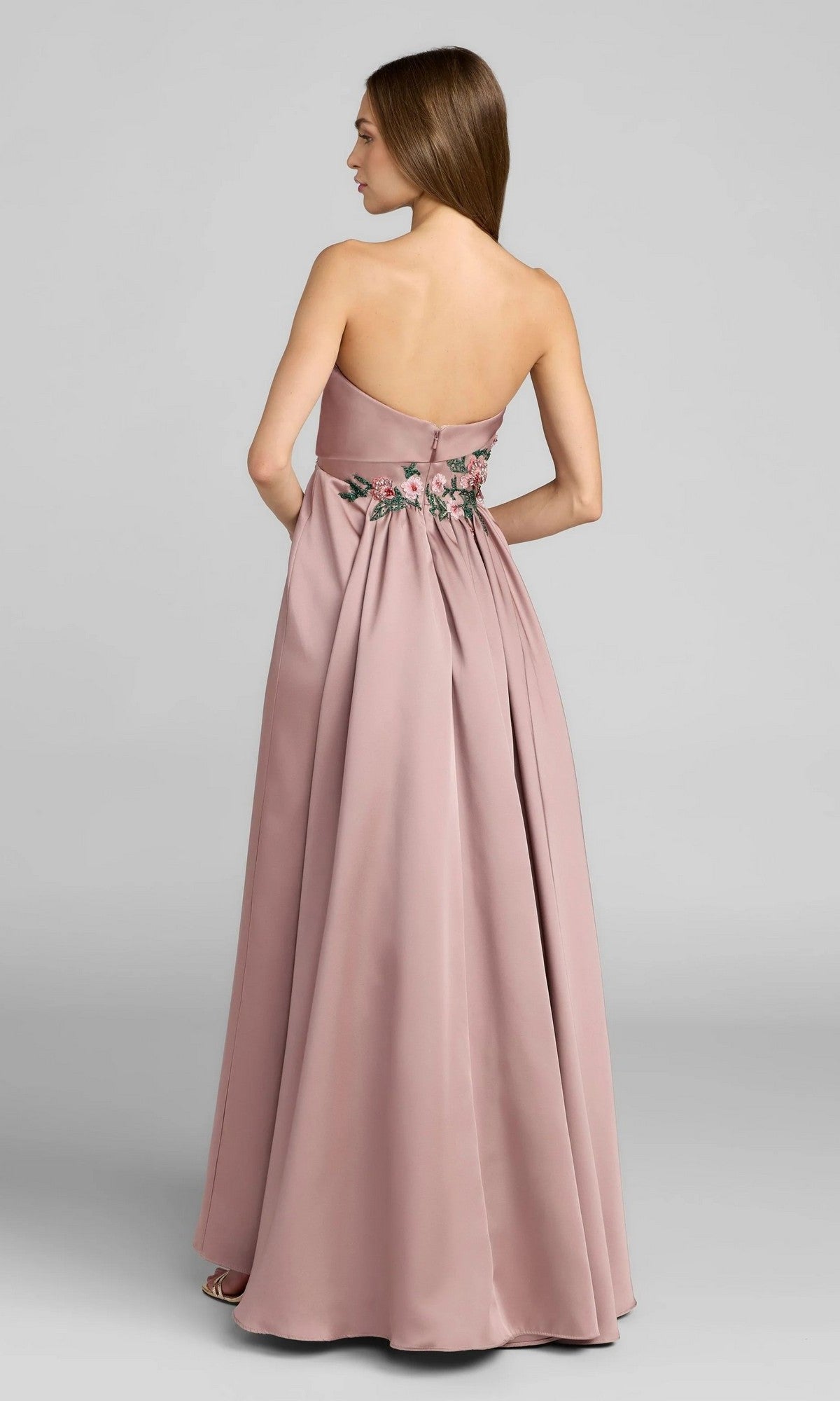 A woman stands facing away in the Mac Duggal 11894—a strapless, floor-length dusty pink gown with floral embroidery at the waist, capturing a timeless elegance characteristic of Mac Duggal’s signature formal style.