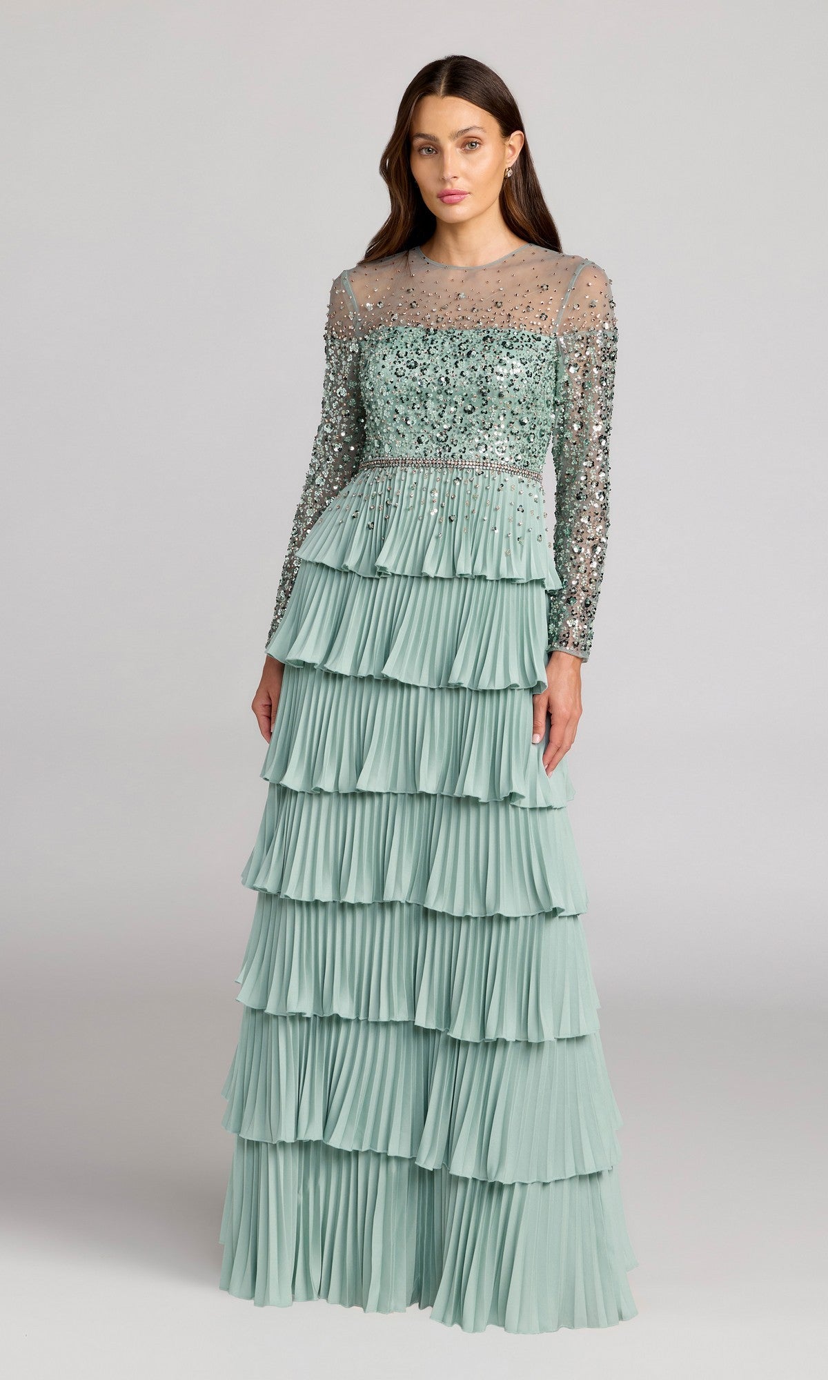 A woman stands wearing the Mac Duggal 11903 long formal dress in sage, featuring a beaded bodice, sheer neckline, long sleeves, and a skirt with multiple pleated ruffle tiers.