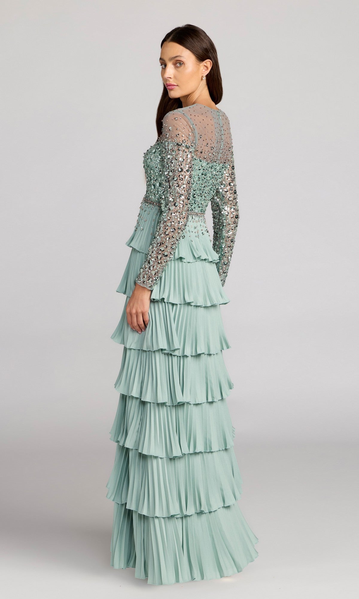 A woman stands against a plain light background wearing the Mac Duggal 11903 long formal dress in sage green, featuring pleated tiers, a sheer sequined bodice, and long embellished sleeves.