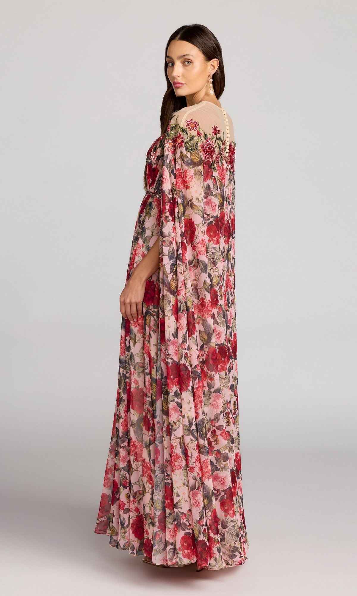 A woman stands sideways against a plain background, looking at the camera while wearing the Mac Duggal 11907 long formal dress—a loose pink floral chiffon gown with sheer detailed shoulders.