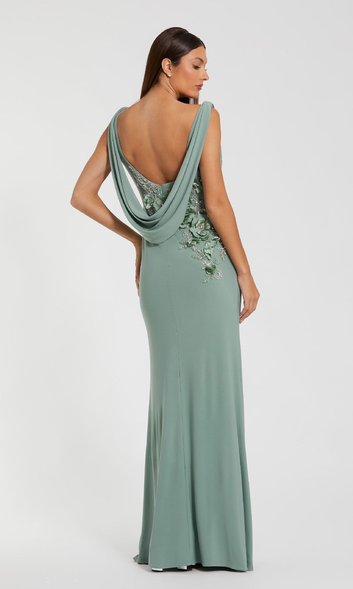 A woman models the Mac Duggal 11910 Cowl-Neck Long Formal Dress in sage green, featuring a floor-length silhouette, draped open back, and floral embroidery, against a plain light background.