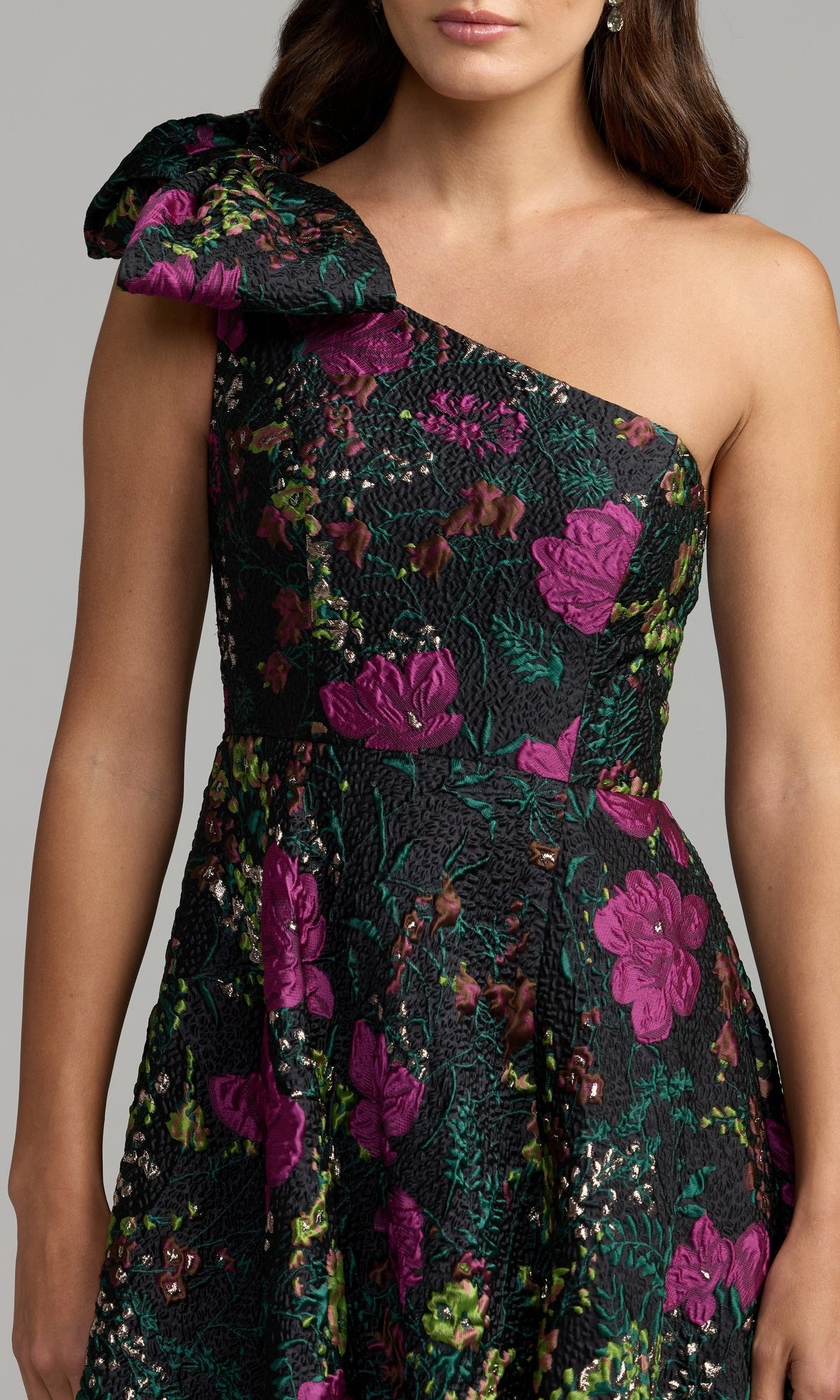 A woman wearing the Mac Duggal 11919 One-Shoulder Floral Formal Gown—a striking black dress featuring textured purple floral patterns and elegant green leafy accents.