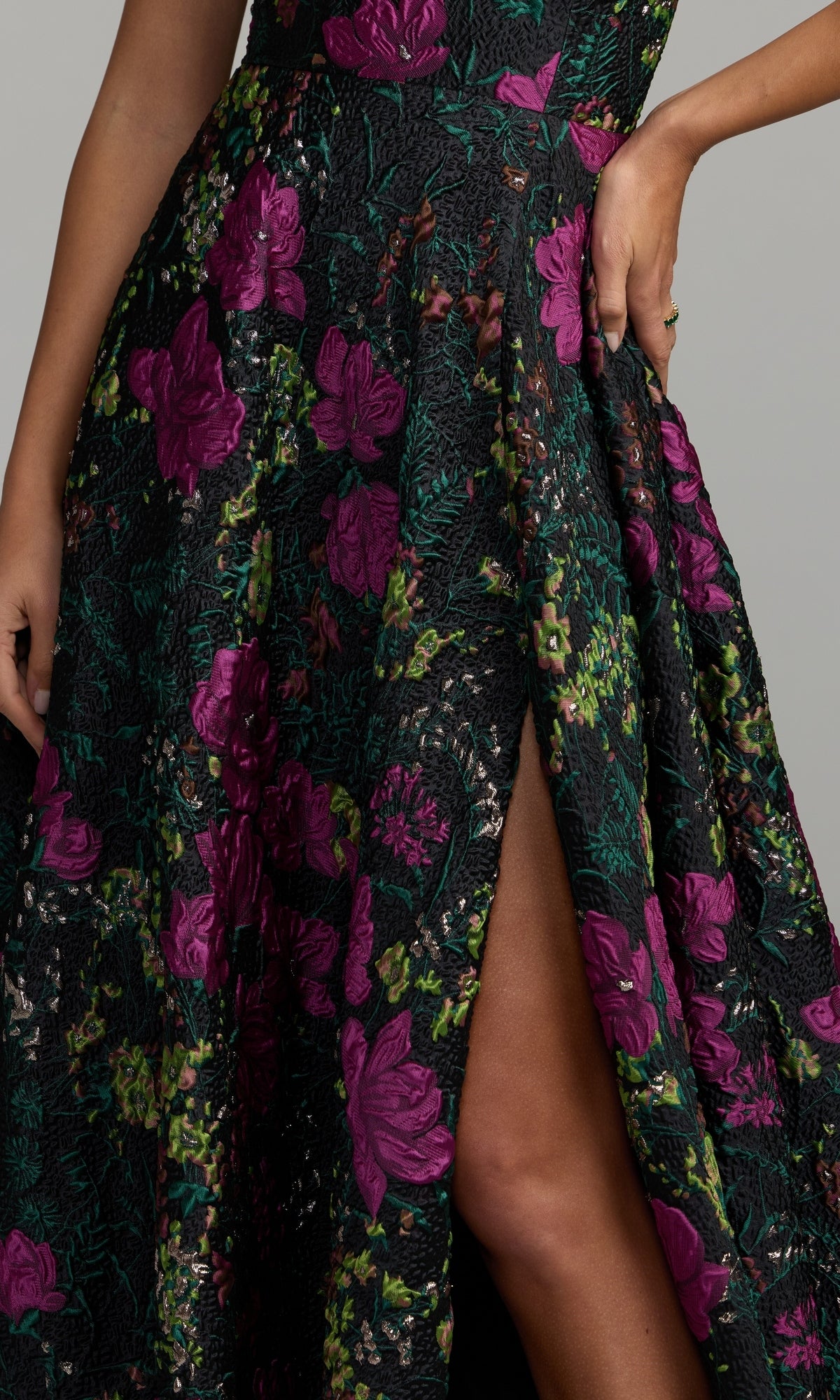 Wearing the One-Shoulder Floral Formal Gown Mac Duggal 11919—an elegant black dress detailed with purple floral patterns and a thigh-high slit—the person stands confidently with one hand on their hip, showcasing a stunning choice for prom or any formal occasion.