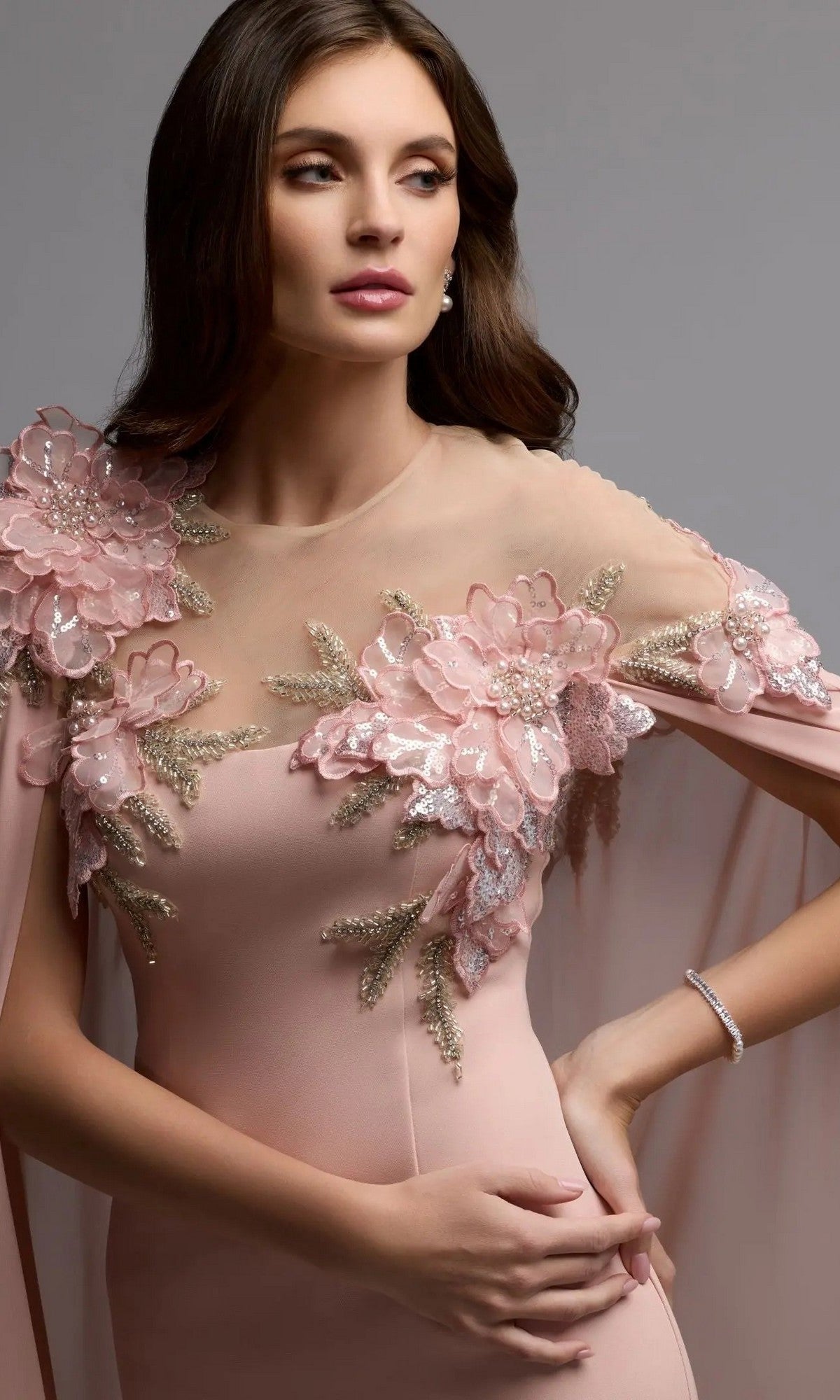 A woman in the Mac Duggal 11942 long formal dress—with a pink hue, sheer neckline, 3D floral appliques, and cape sleeves—stands against a plain gray background.