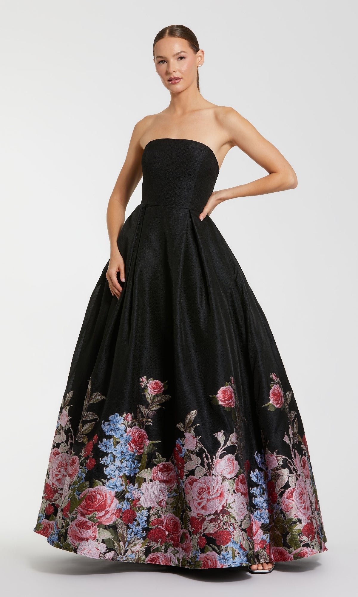 A woman stands against a plain background wearing the Mac Duggal 11970 Strapless Floral Prom Ball Gown, a long black dress with a full skirt adorned with pink and blue floral patterns near the hem.