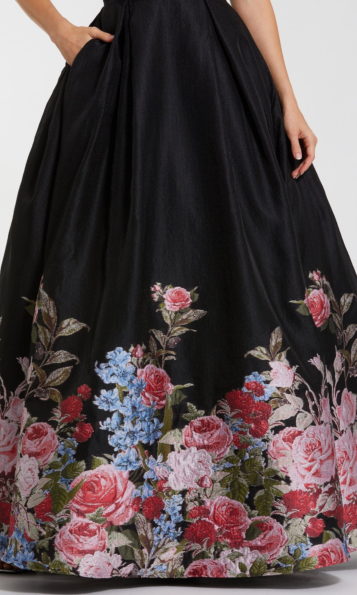 A person in the Mac Duggal 11970 Strapless Floral Prom Ball Gown—a long black dress with colorful roses and blue floral patterns along the hem.