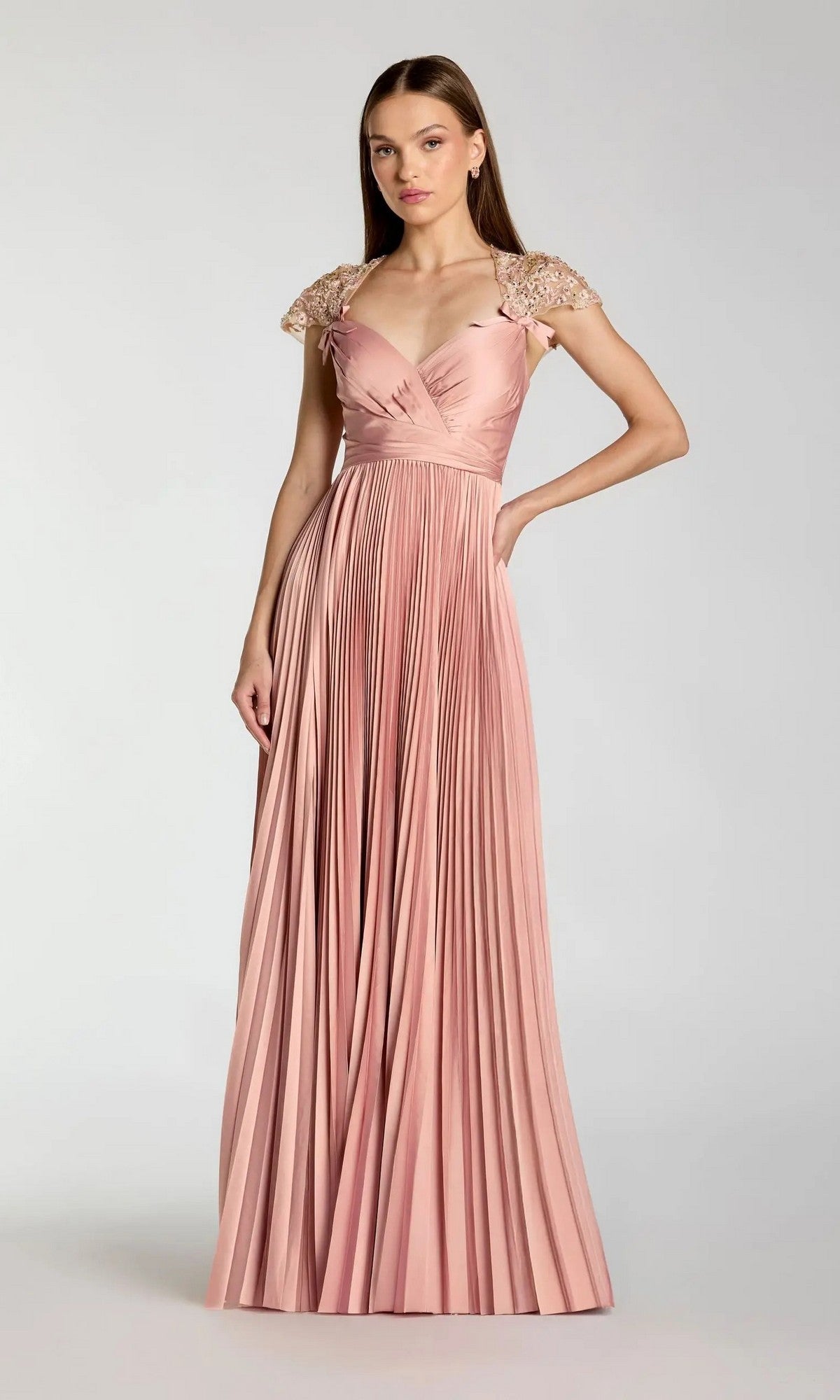 A woman wearing the Mac Duggal 11987 long formal dress, featuring floor-length pleats and embellished cap sleeves in pink, stands against a plain light gray background.
