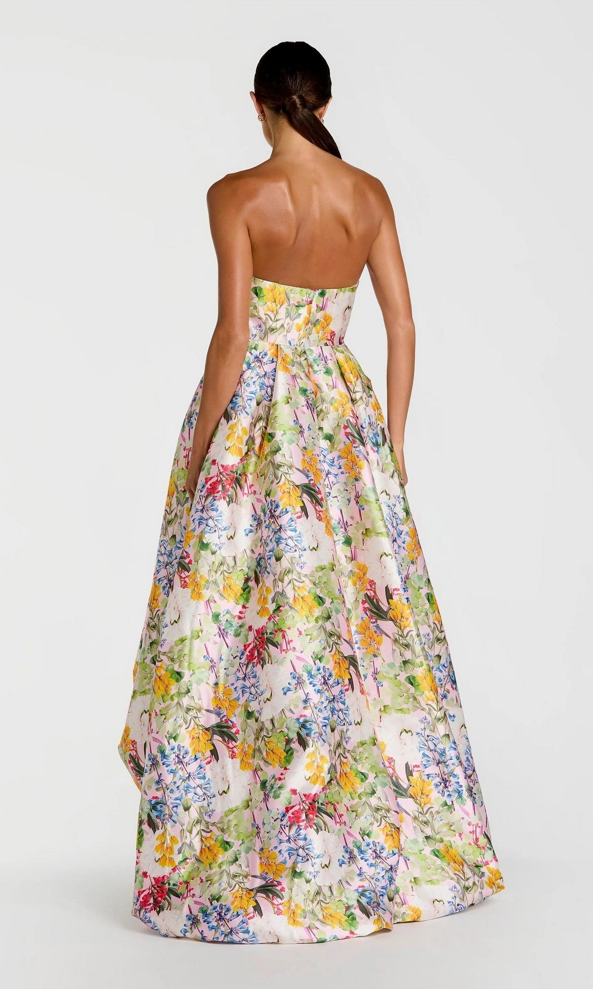 A woman faces away, wearing the Mac Duggal 11991 long formal dress—strapless with a vibrant floral print on a light background and featuring a dramatic high-low bubble hem.