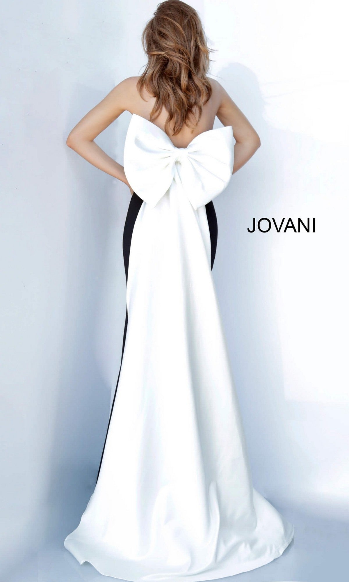 A woman is seen from behind wearing the Jovani 12020, a strapless black satin mermaid formal dress with an oversized back bow and a long white train. The word “JOVANI” appears on the wall.