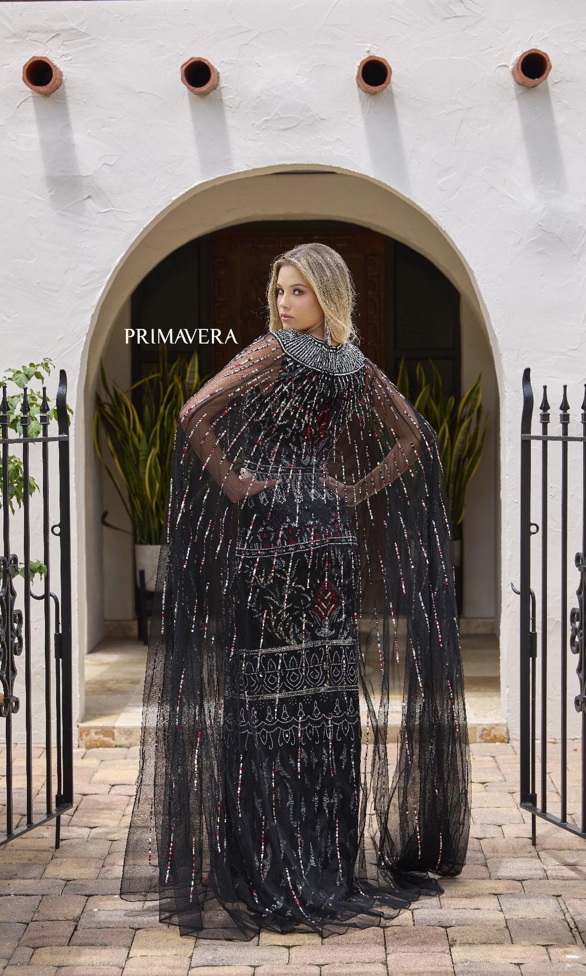 A woman wears the Primavera 12172 long formal dress with a sheer, beaded cape, standing with her back to an arched doorway framed by black gates and potted plants.