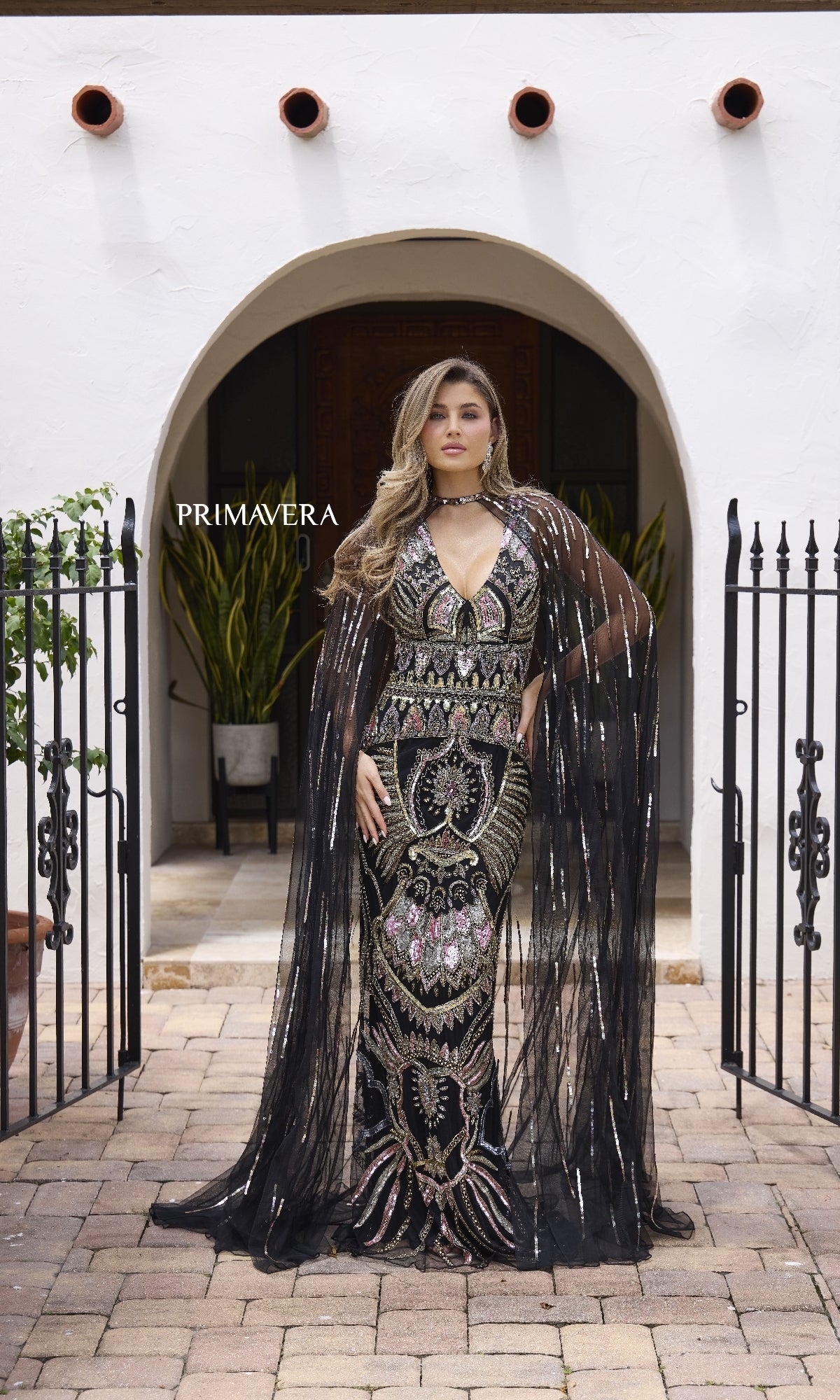 A woman stands before an arched doorway, wearing the Primavera 12177 long formal dress—black and gold sequined, sleeveless, with a sheer floor-length cape—framed by iron gates and potted plants.
