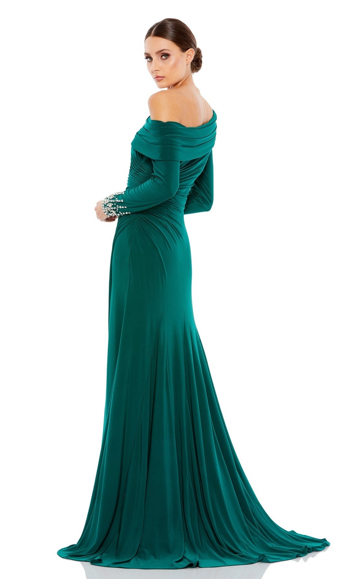 A woman in a Mac Duggal 12231 one-shoulder long formal dress stands against a white background, looking over her shoulder; the emerald green gown features an elegant A-line silhouette and beaded cuff details.