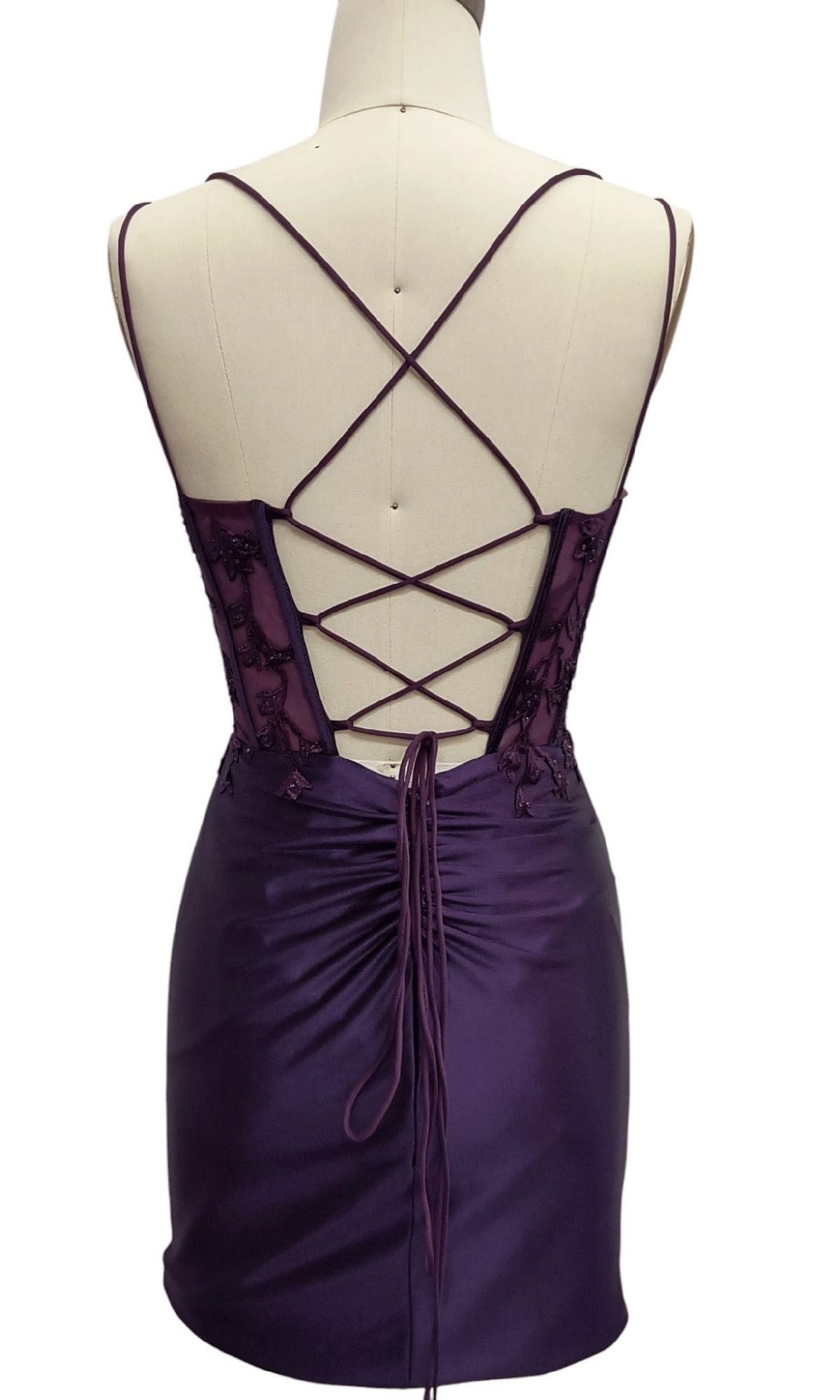 Back view of mannequin in Short Purple Hoco Dress: 12415, a satin homecoming dress with thin crisscross straps, lace-up back detail, and ruched center gathering.