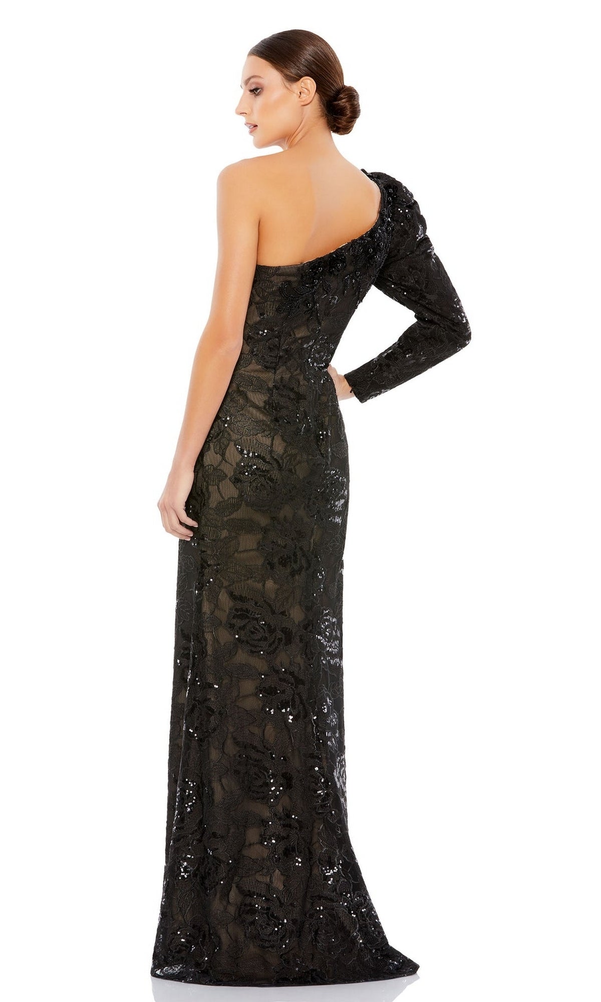A woman stands facing away, wearing the Long Formal Dress 12444 by Mac Duggal—a black, lace, one-shoulder gown embellished with sequins and floral patterns. Her hair is styled in a low bun.