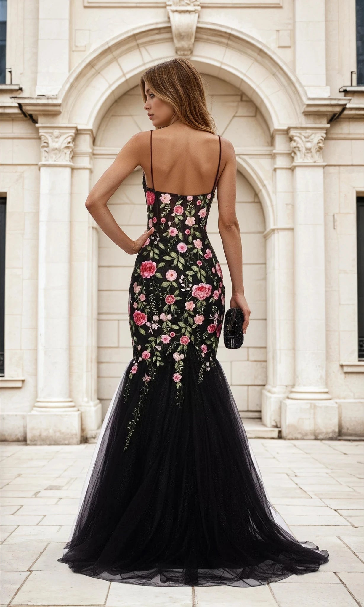 Woman in a Floral-Print Black Glitter Mermaid Prom Dress L488 holds a black clutch, standing before a white stone building with elegant arched details.