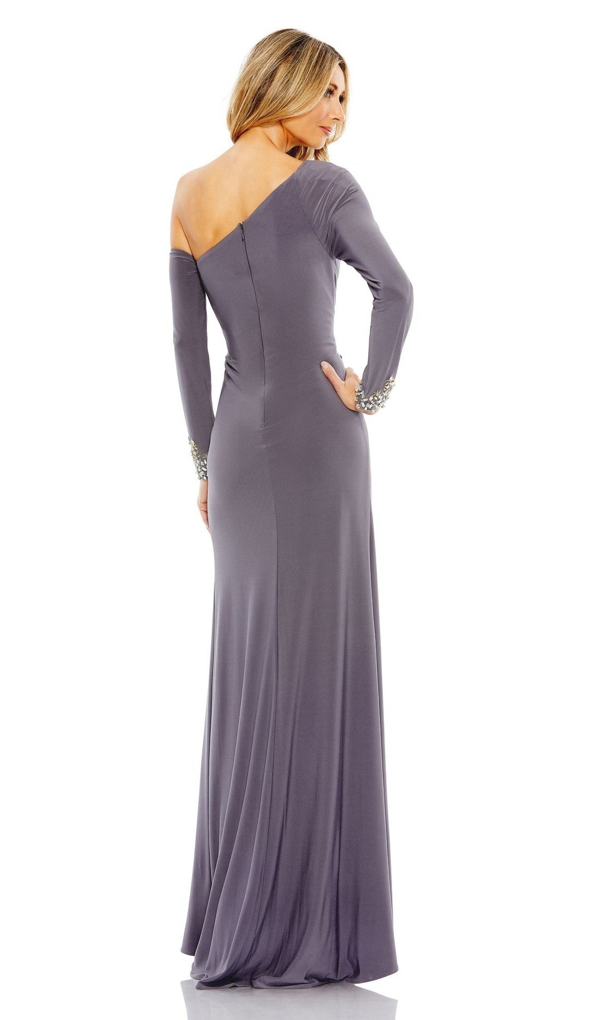A woman stands facing away, wearing the Mac Duggal Long Formal Dress 12489—a fitted, one-shoulder gray gown with long sleeves and beaded cuffs.