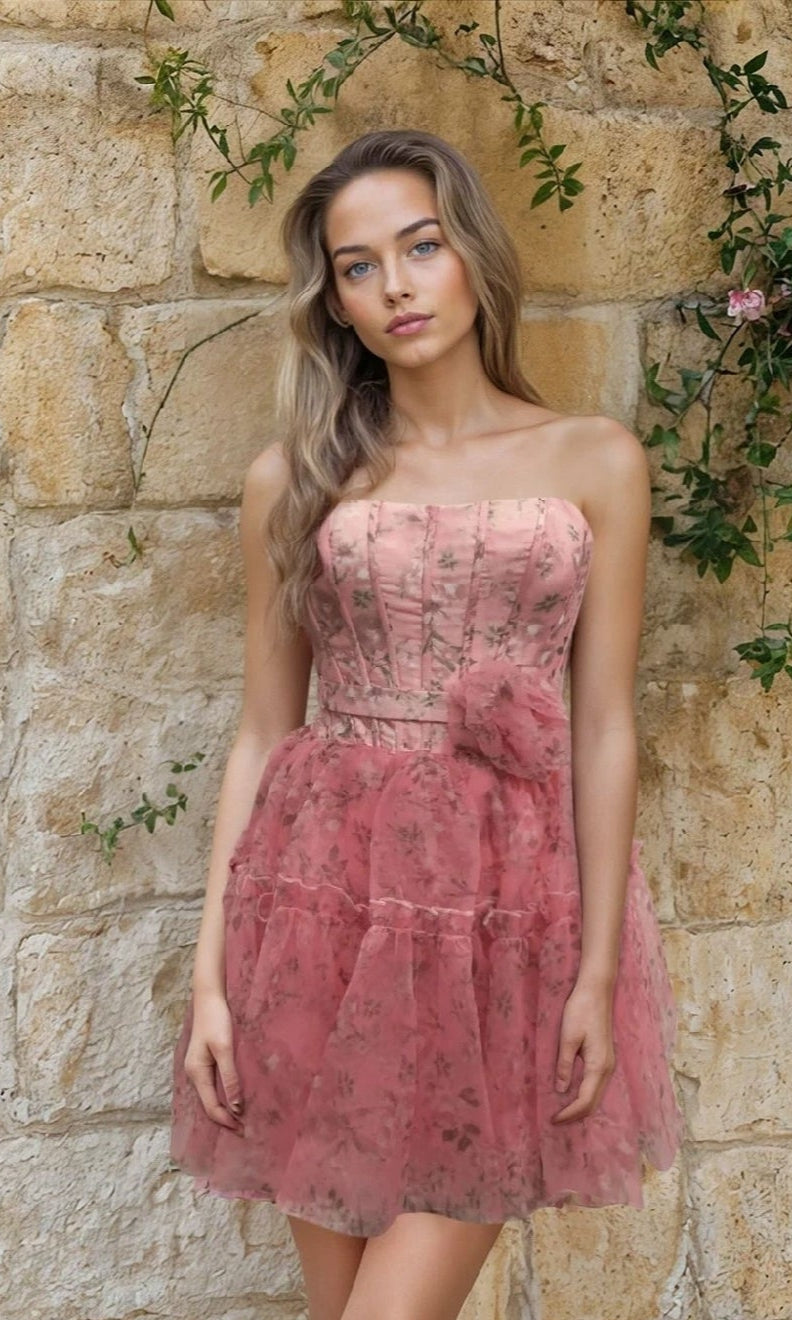 A woman stands before a stone wall in the Strapless Short Pink Hoco Dress: 12603, featuring sheer layers, a waist bow, and a floral print design.