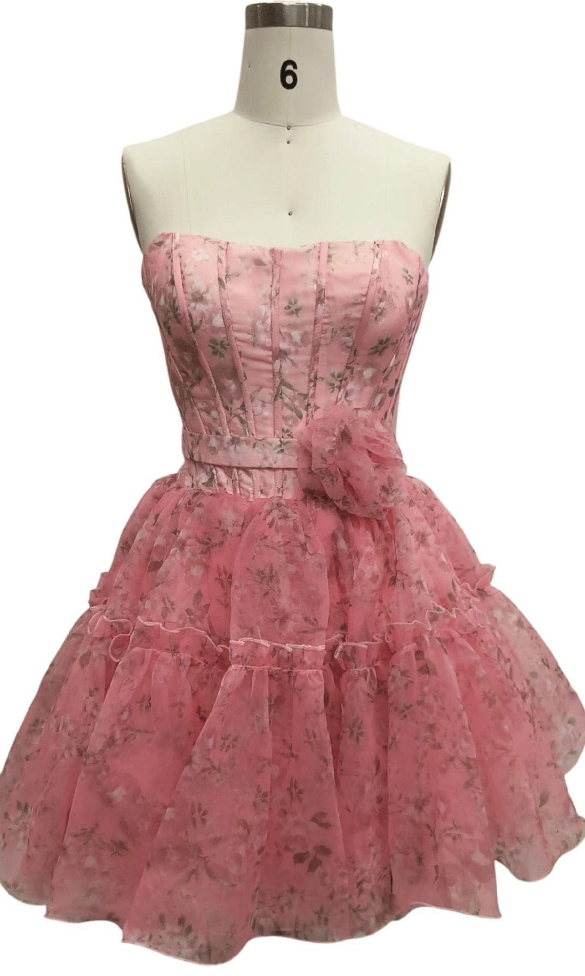 The Strapless Short Pink Hoco Dress: 12603 features a floral print, corset-style bodice, layered ruffled skirt, and waist flower detail. Displayed on a dress form, it's the perfect pink party mini dress.