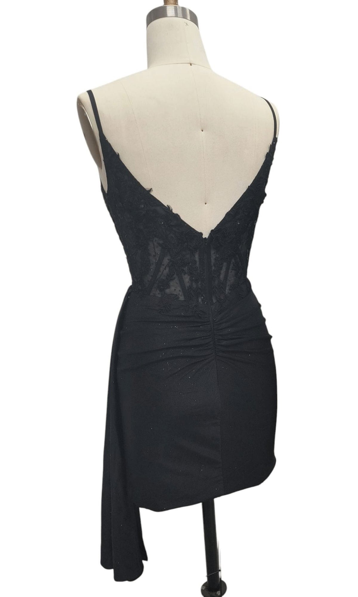 The Side-Drape Short Hoco Dress: 12658 is a black sleeveless homecoming dress with lace bodice detailing and an asymmetrical draped skirt, displayed from the back on a dress form.