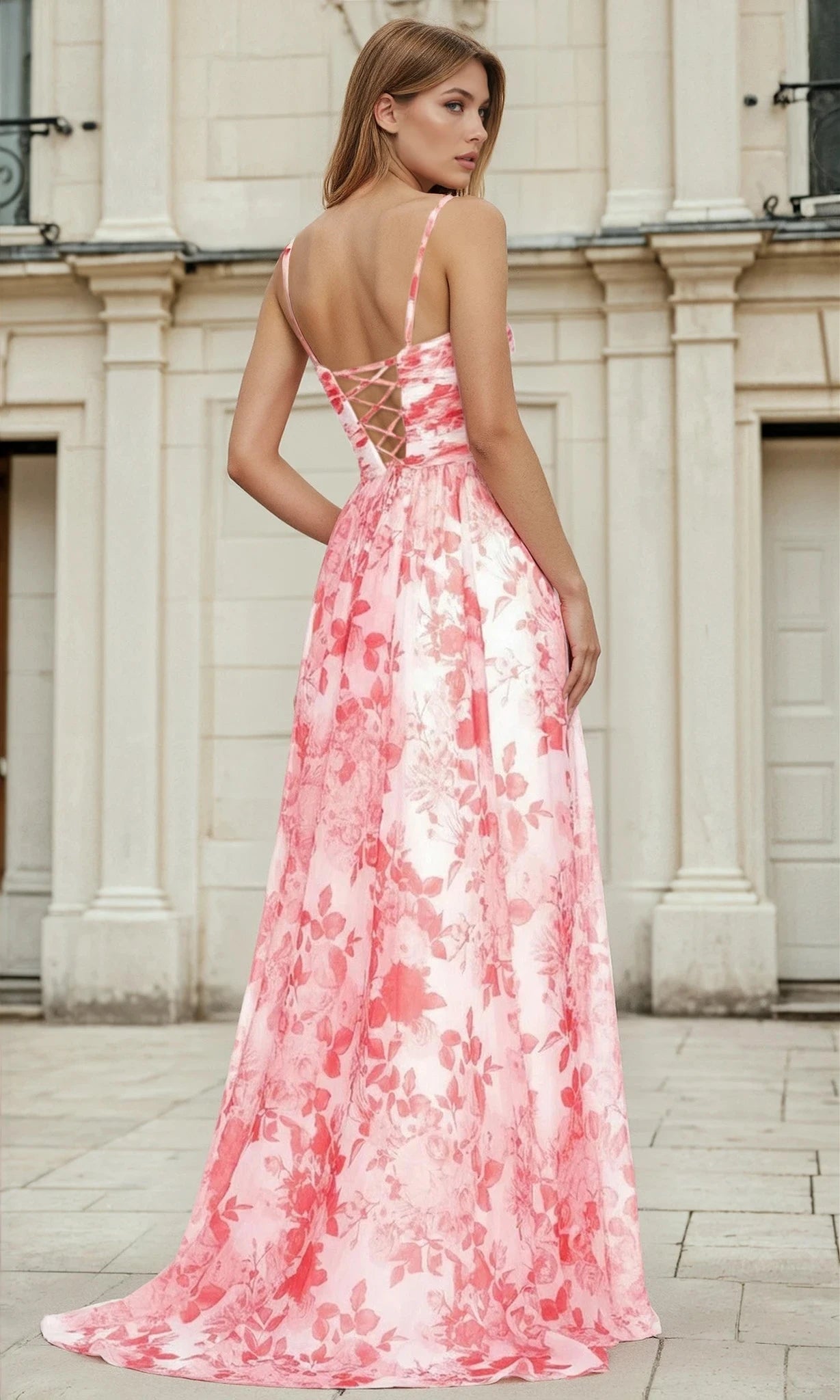 A woman stands outdoors in front of a light-colored building, looking over her shoulder while wearing the Long A-Line Floral Prom Dress L693, which features a pink floral pattern, sweetheart neckline, and lace-up back.