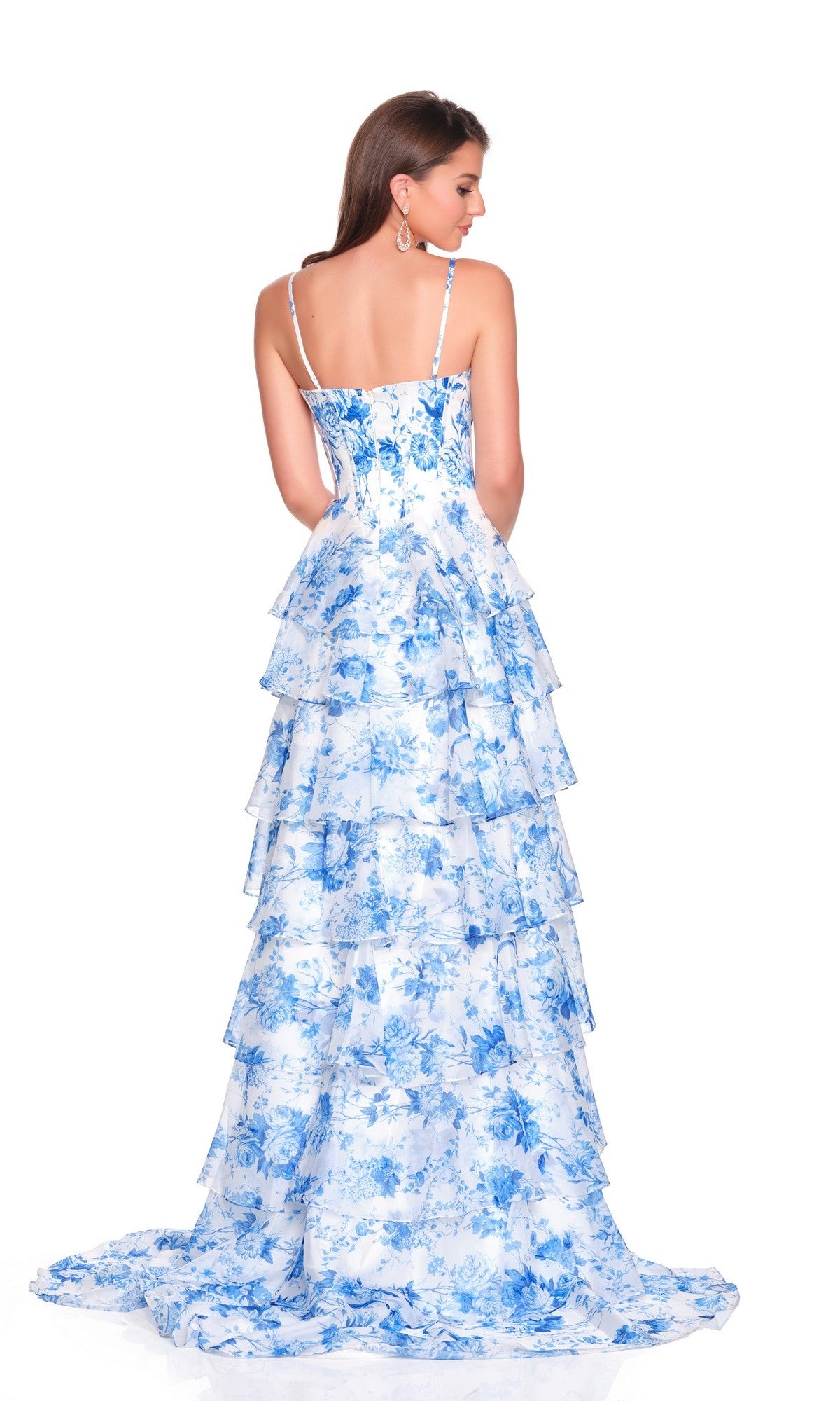 A woman stands with her back to the camera against a plain white background, wearing the Long Prom Dress: Dave and Johnny 12720—a long, tiered, white chiffon A-line skirt with blue floral patterns and a satin corset bodice.