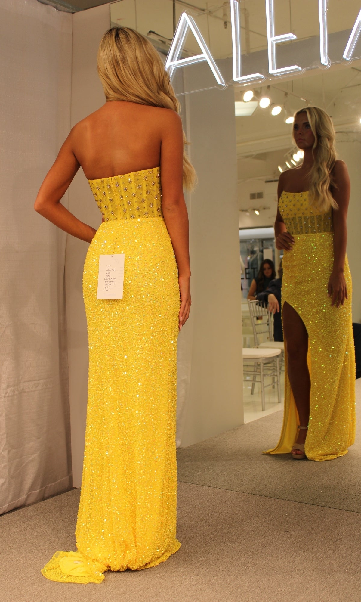 A woman wearing the Aleta Strapless Beaded Designer Prom Dress 1278, featuring a yellow sparkly design and thigh-high slit, stands before a mirror showing both her front and back reflections.