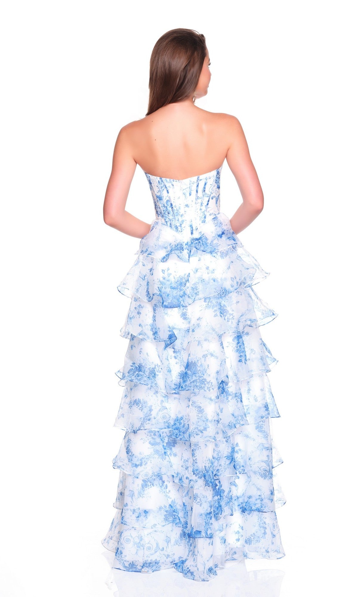 A woman facing away wears the Dave and Johnny 12936 long prom dress—a strapless, floor-length white gown with blue floral patterns and layered ruffles—standing against a white backdrop.