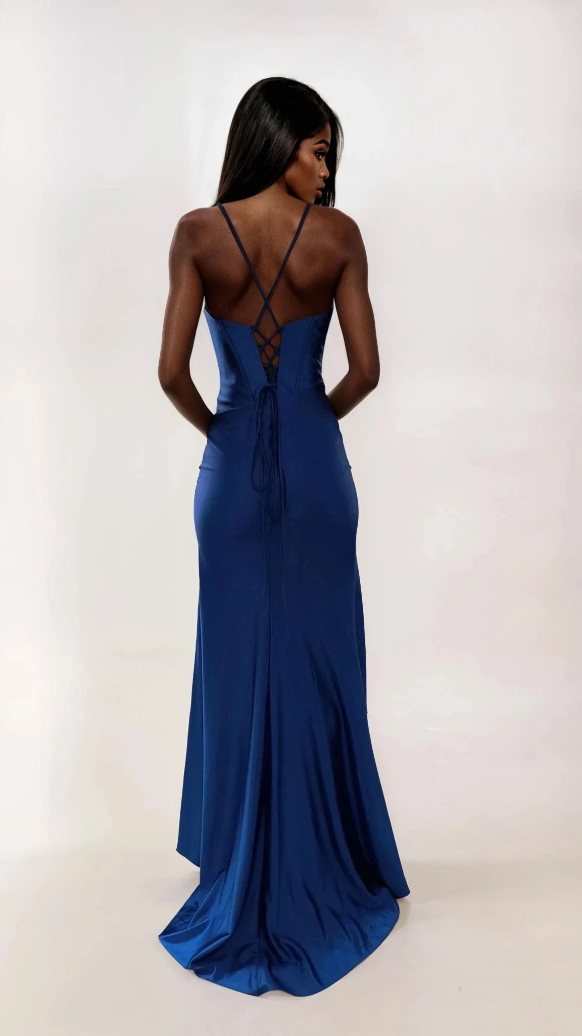 A woman faces away in the Dave and Johnny 12954 long prom dress, featuring a deep blue fitted design with crisscross straps, a lace-up corset bodice, and a floor-length satin skirt set against a plain light background.