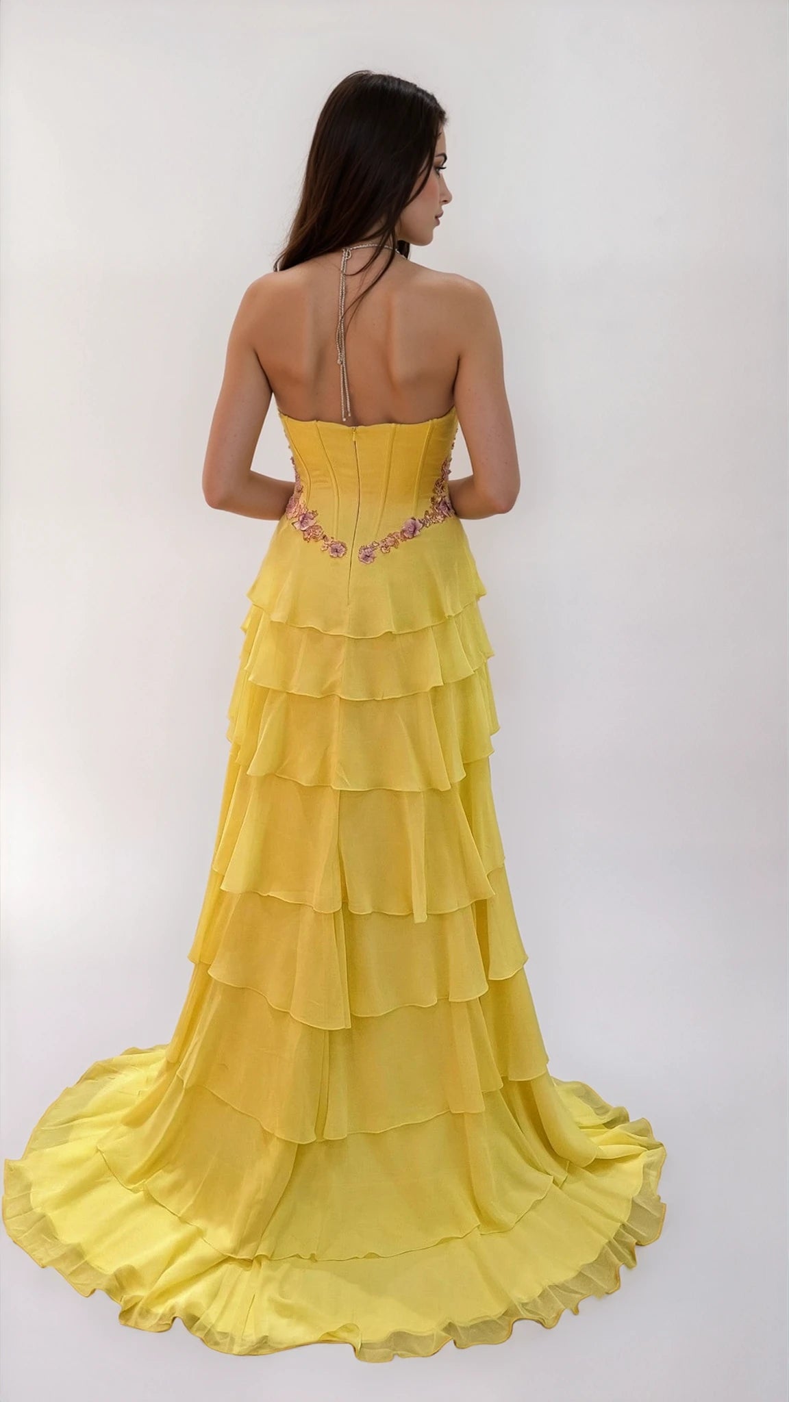 A woman faces away, wearing the Dave & Johnny 12966 long tiered A-line prom dress with a strapless yellow design and floral embroidery at the waist, set against a plain white background.