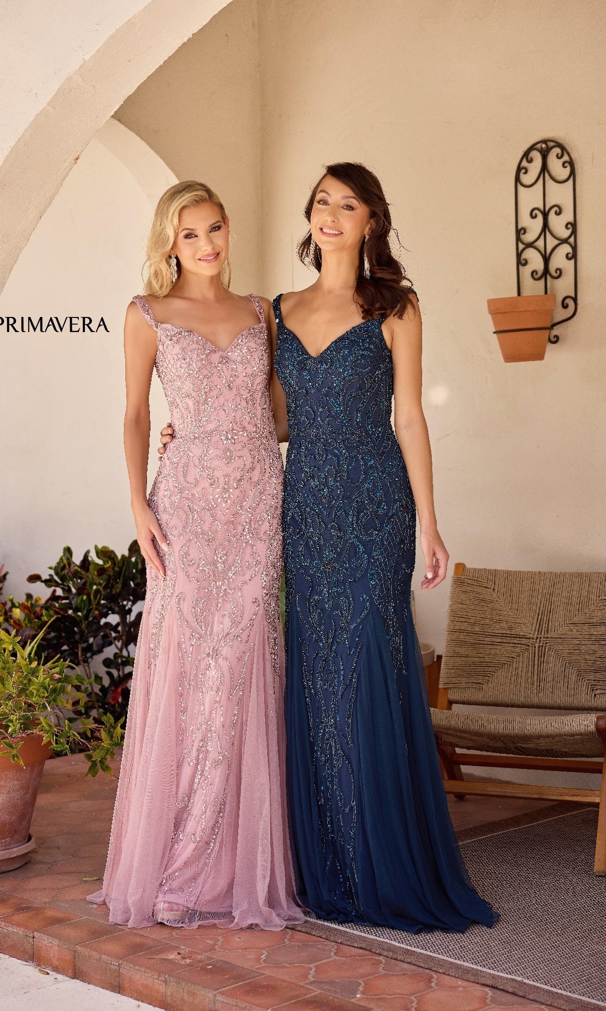 Two women pose side by side on a simply decorated patio, each wearing the Primavera 13143 long formal dress—one in pale pink, the other in dark blue—both featuring sparkly embellishments.