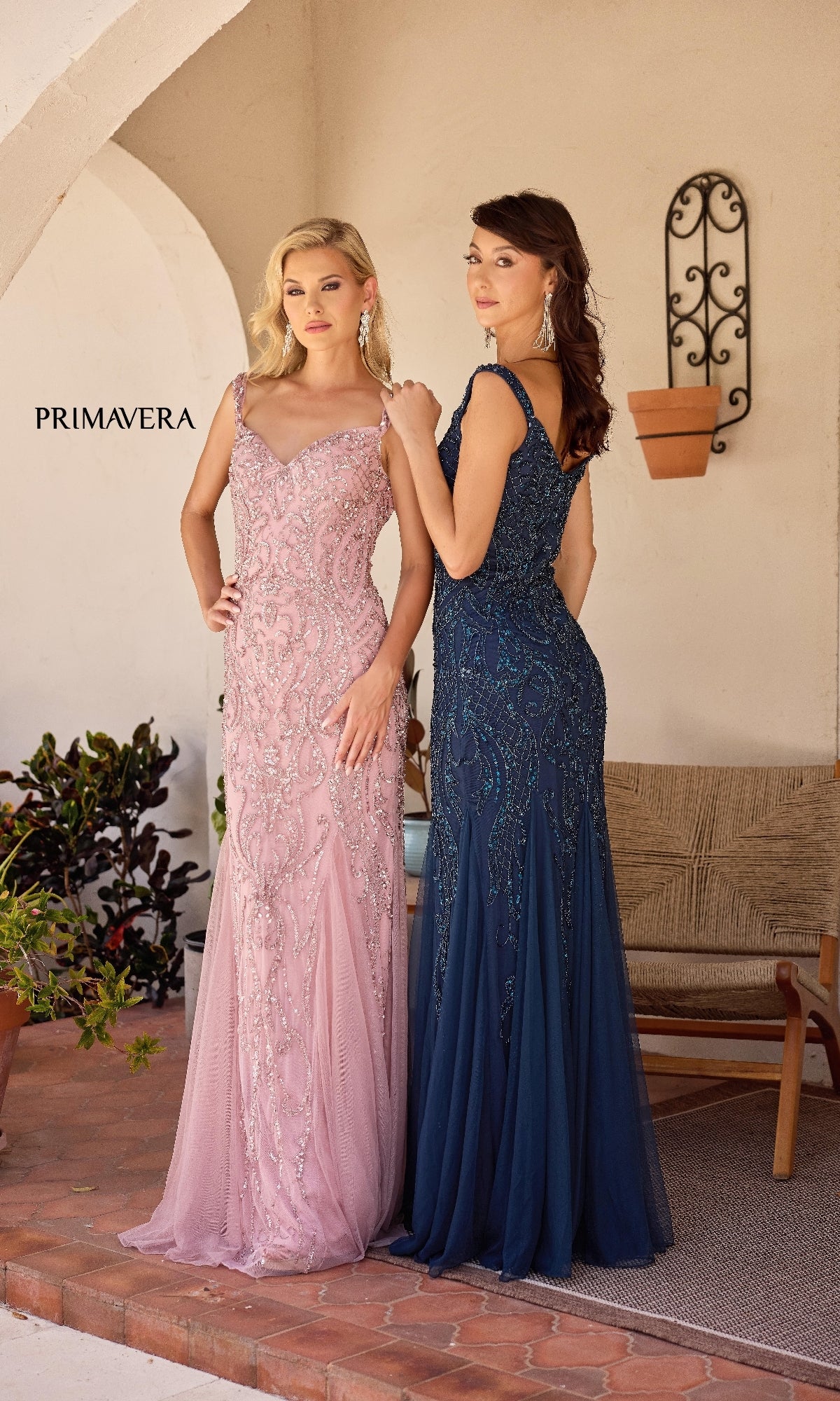 Two women in Primavera 13143 long formal dresses—one pink with a sweetheart neckline, the other navy blue—stand side by side outdoors near a bench and potted plants.