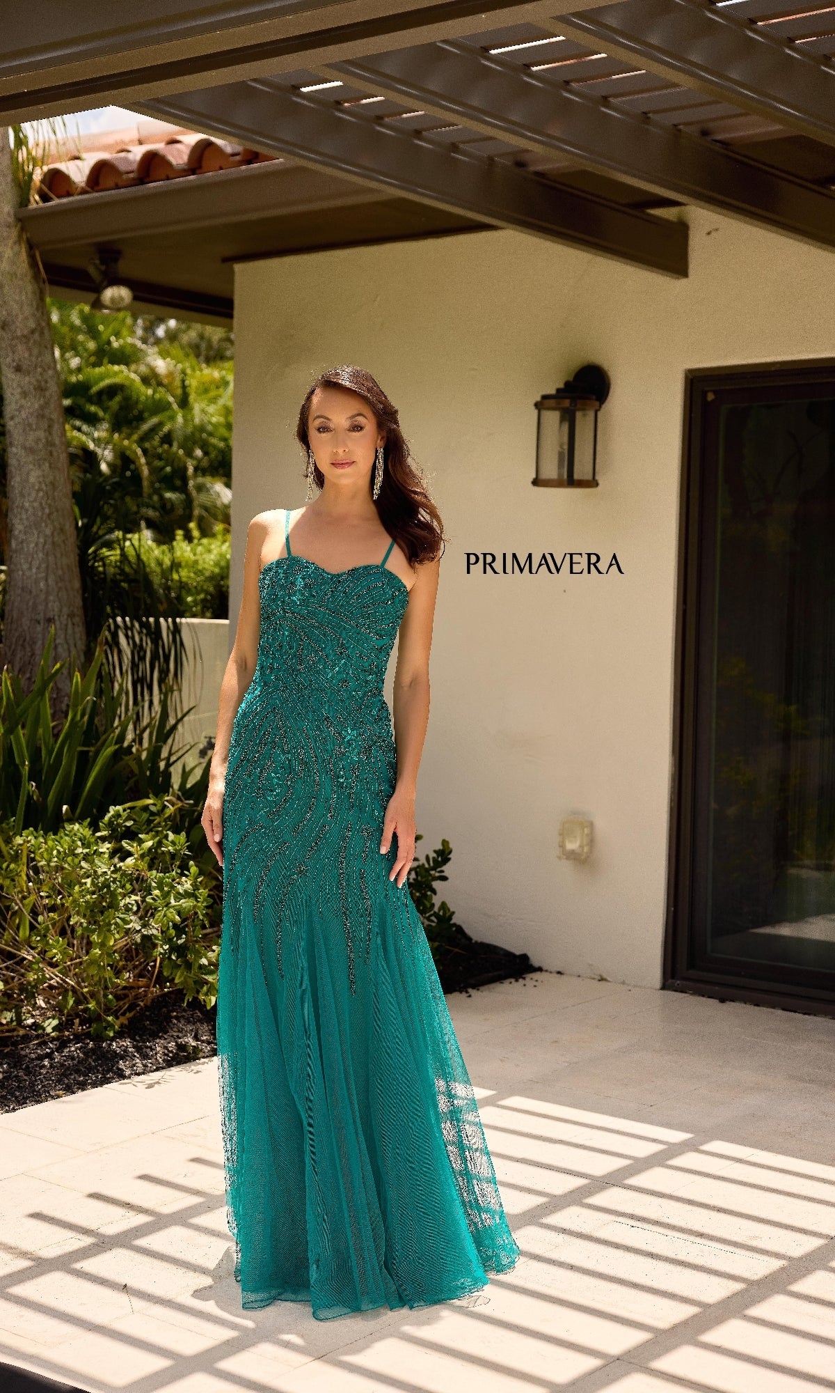 A woman in the Sheer Capelet Long Formal Dress: Primavera 13145 stands outside a white-walled building with a black door, under a covered patio. The word