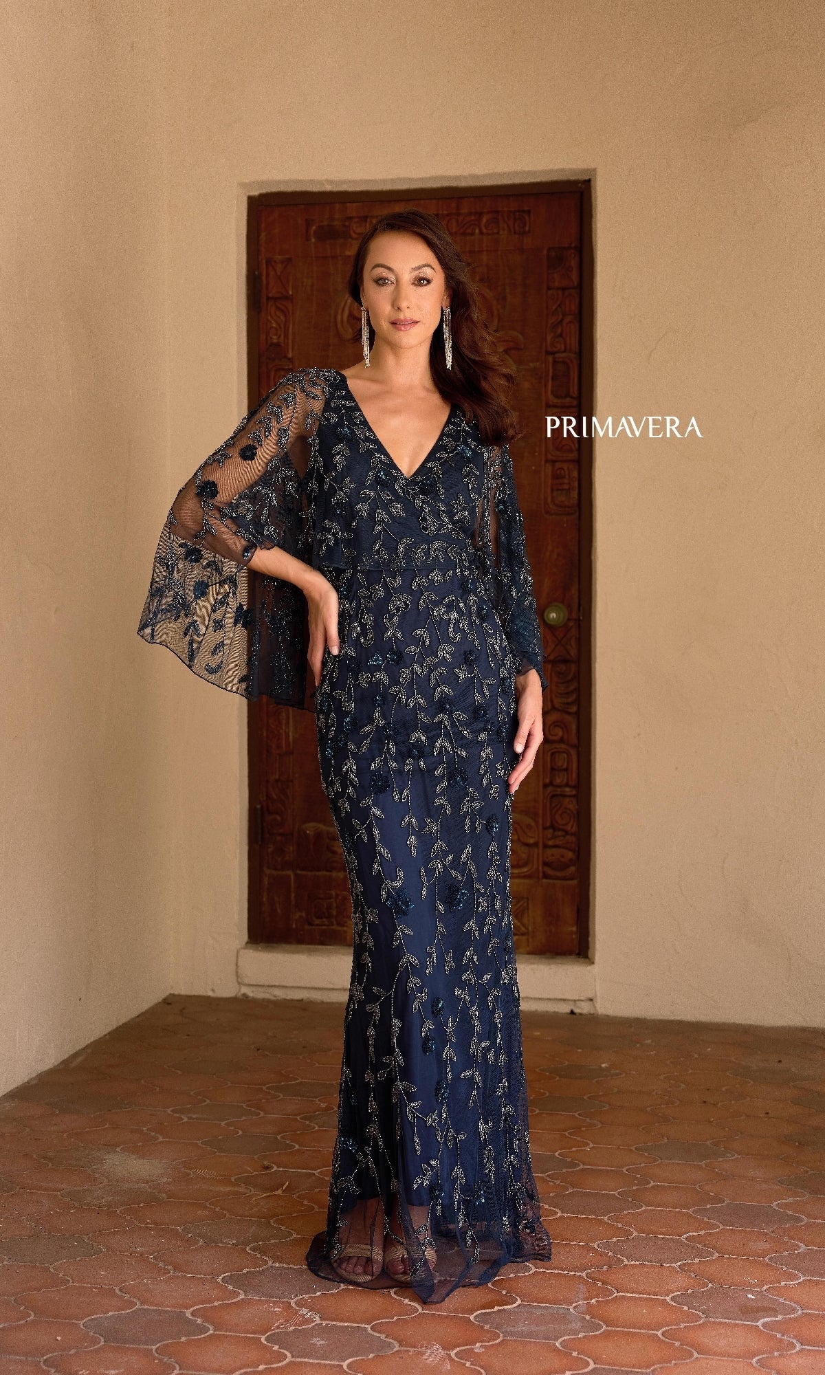 A woman stands in front of a wooden door wearing the Primavera 13149, a navy blue long formal dress featuring silver floral embroidery, beaded details, a v-neckline, and flared sleeves.