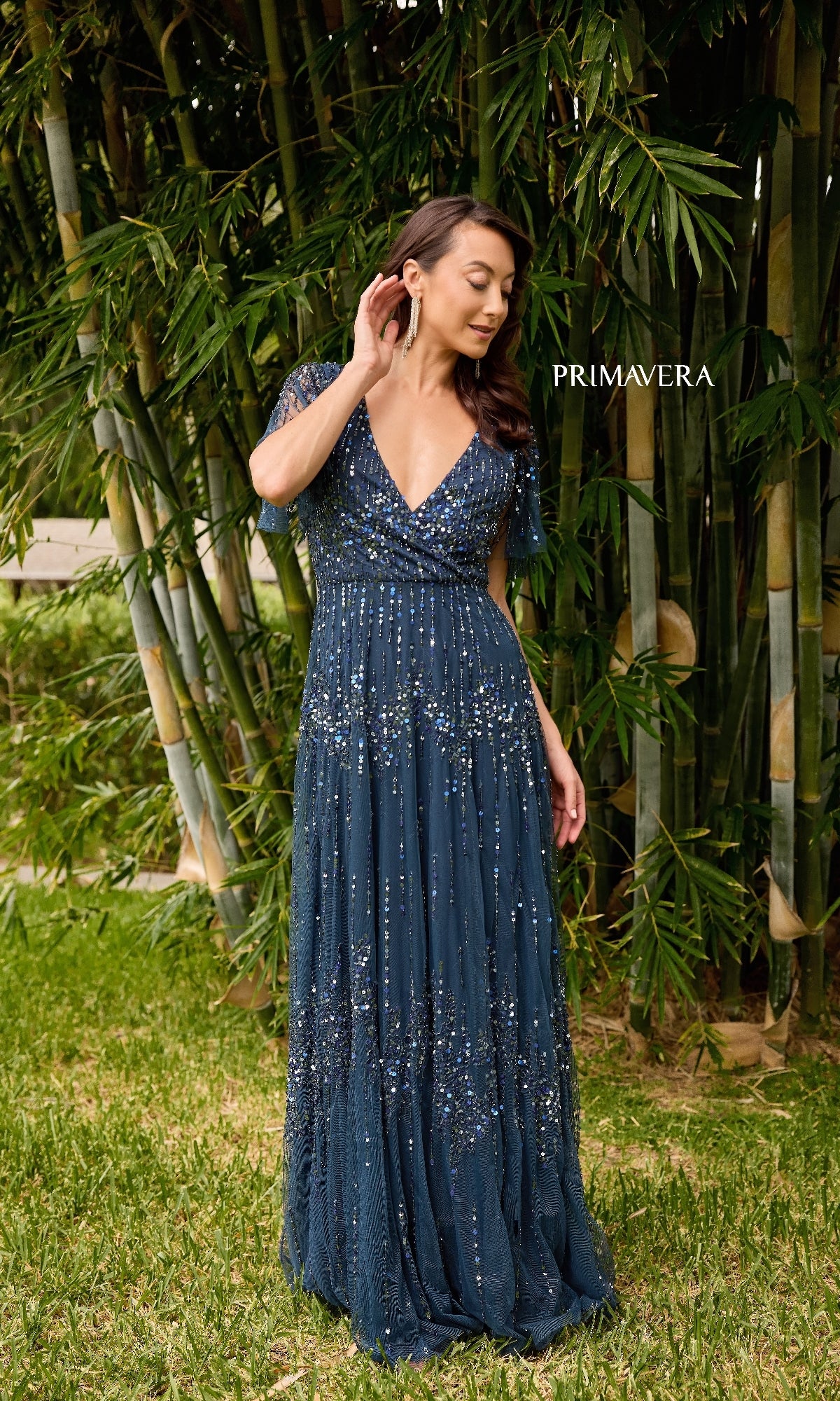 A woman in the Primavera 13153 long formal dress, a deep blue floor-length A-line sequin gown, stands on grass before tall bamboo, touching her earring. The word