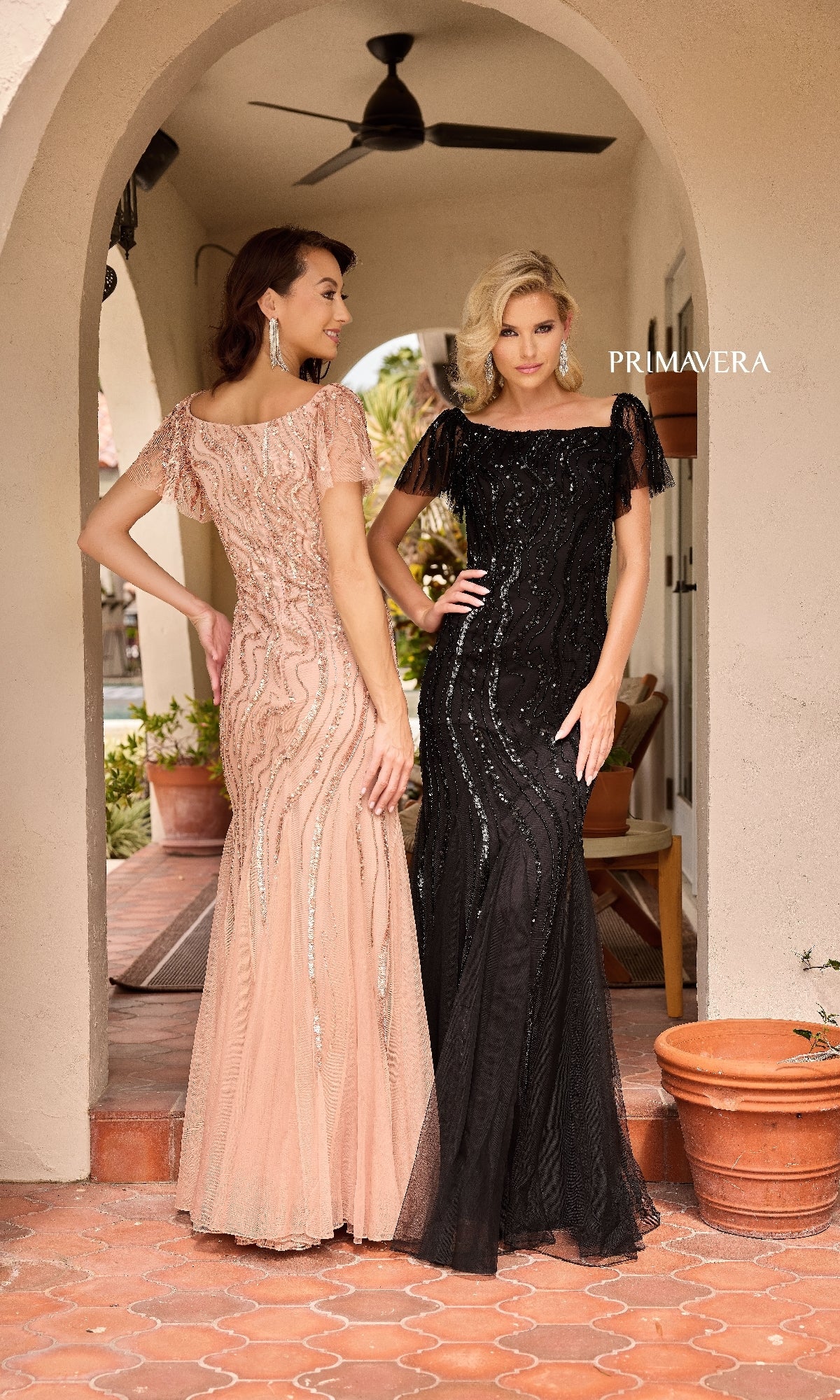 Two women pose on a tiled porch in glamorous Primavera 13155 long formal dresses—one blush pink, the other black—under a ceiling fan near potted plants, radiating elegance in sequined mermaid gowns.