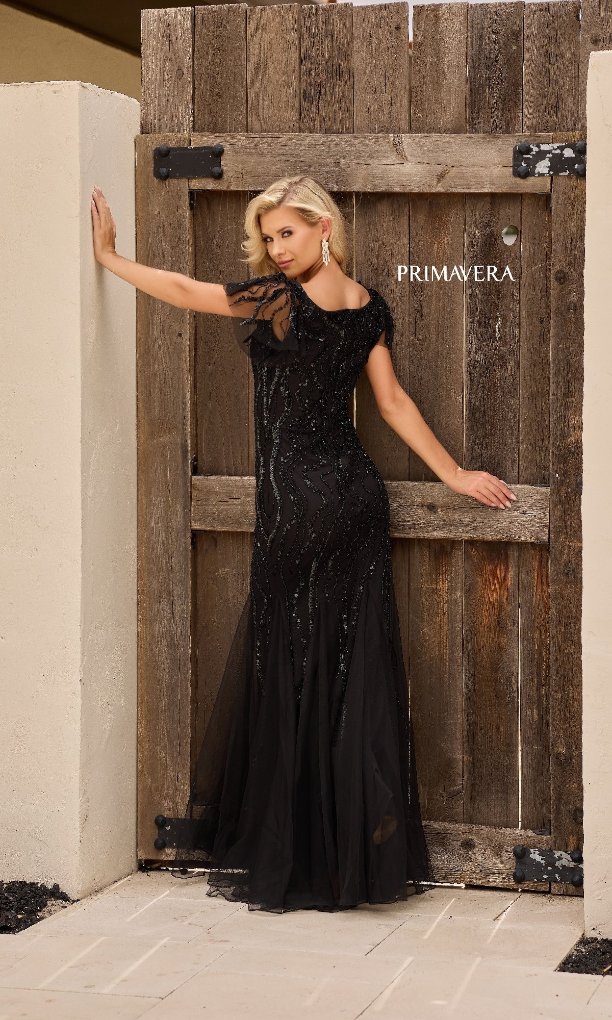 A woman poses by a wooden gate labeled “PRIMAVERA,” wearing the Long Formal Dress: Primavera 13155—a black, floor-length gown with elegant sequin-inspired embellishments, reminiscent of a mermaid dress.