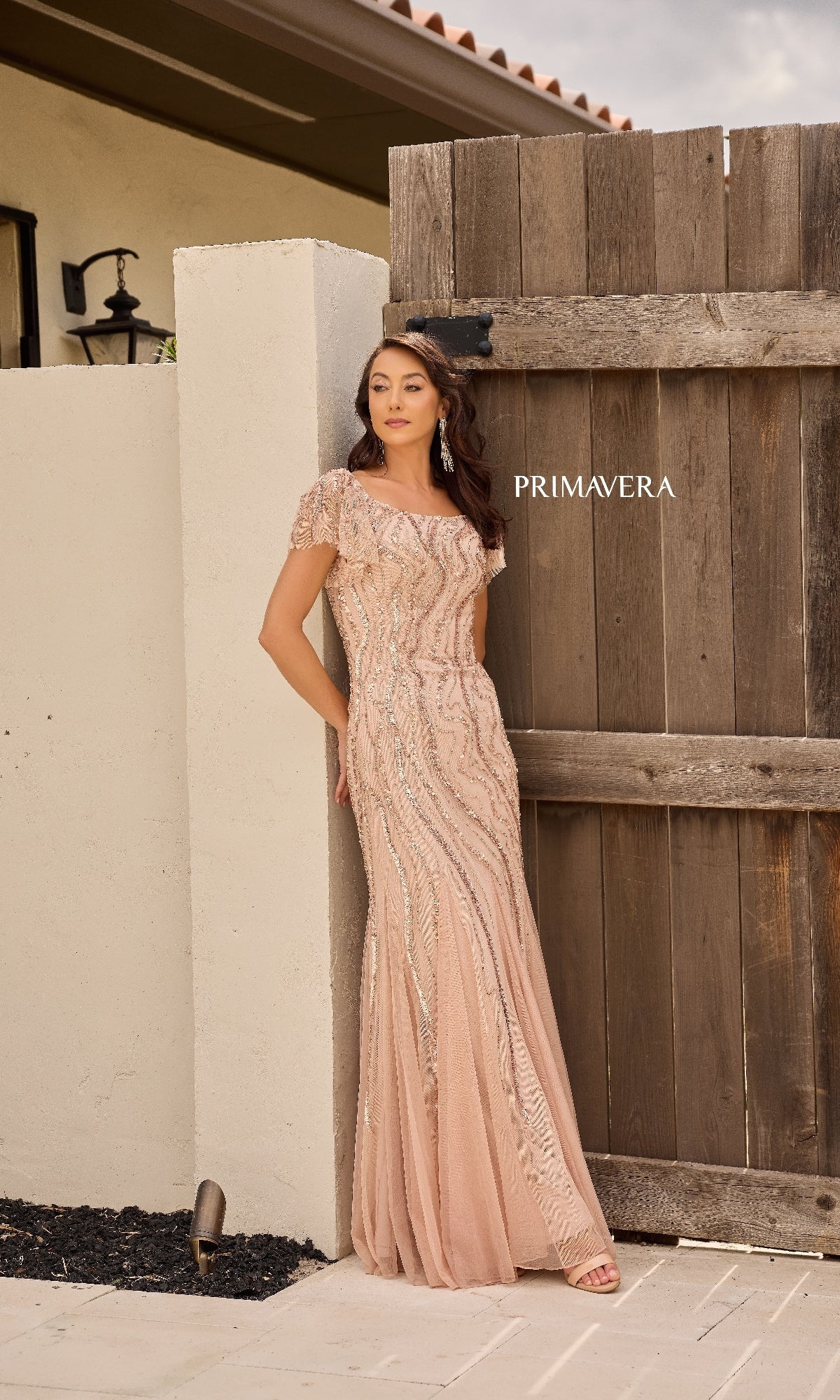 A woman stands outdoors by a wooden gate and white wall, wearing the Primavera 13155—a long, blush-colored formal dress with embellishments and short sleeves.
