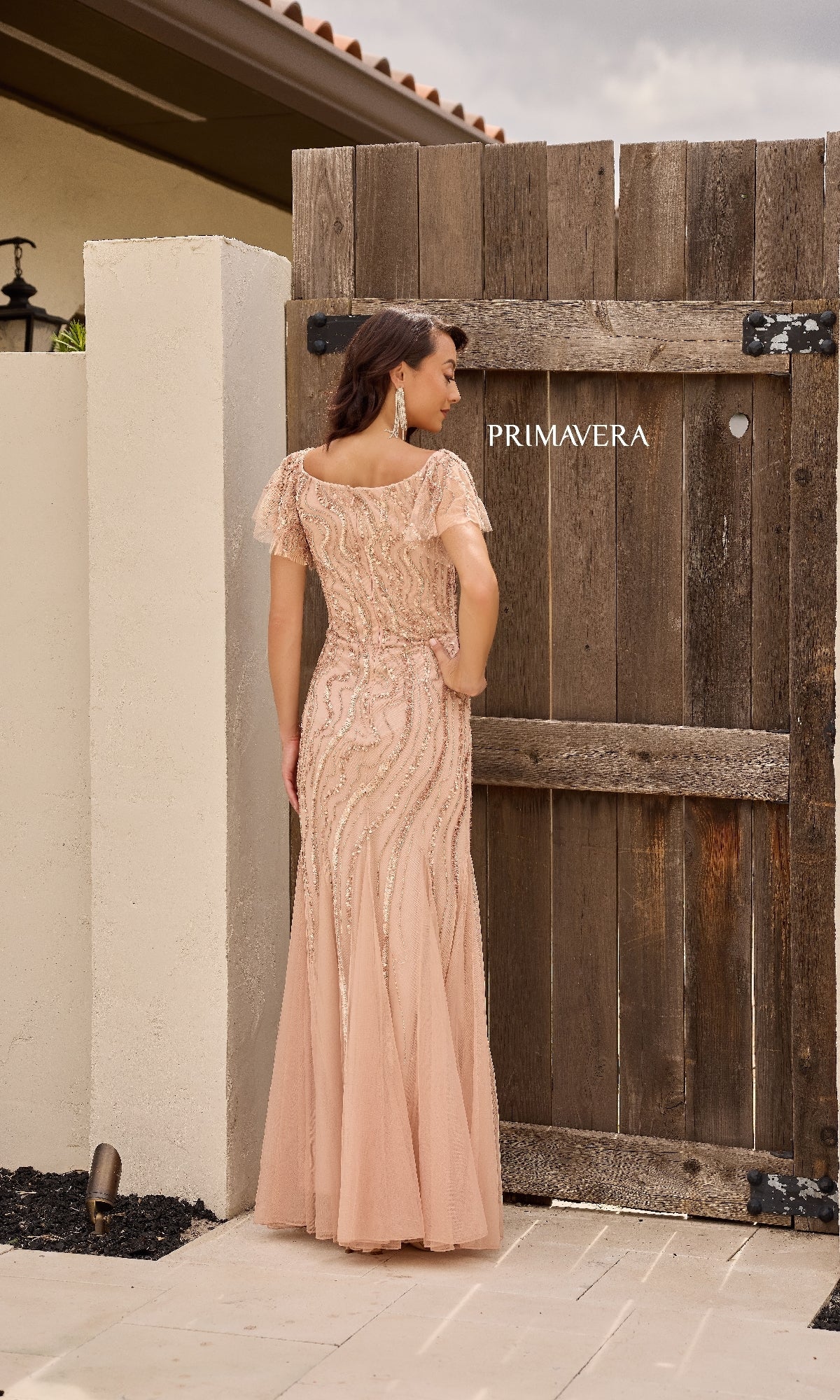 A woman wearing the Primavera 13155 long formal dress with short sleeves and embellishments stands with her back to the camera in front of a rustic wooden gate.